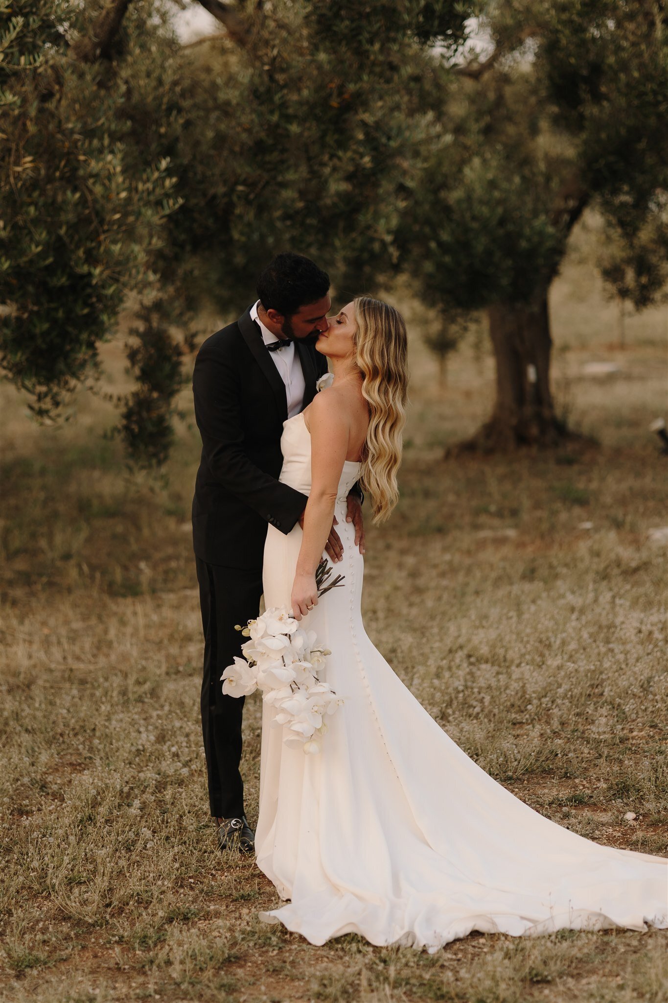 Bride and groom stand in an olive grove at Masseria Moroseta, kissing. The bride holds a orchids bouquet