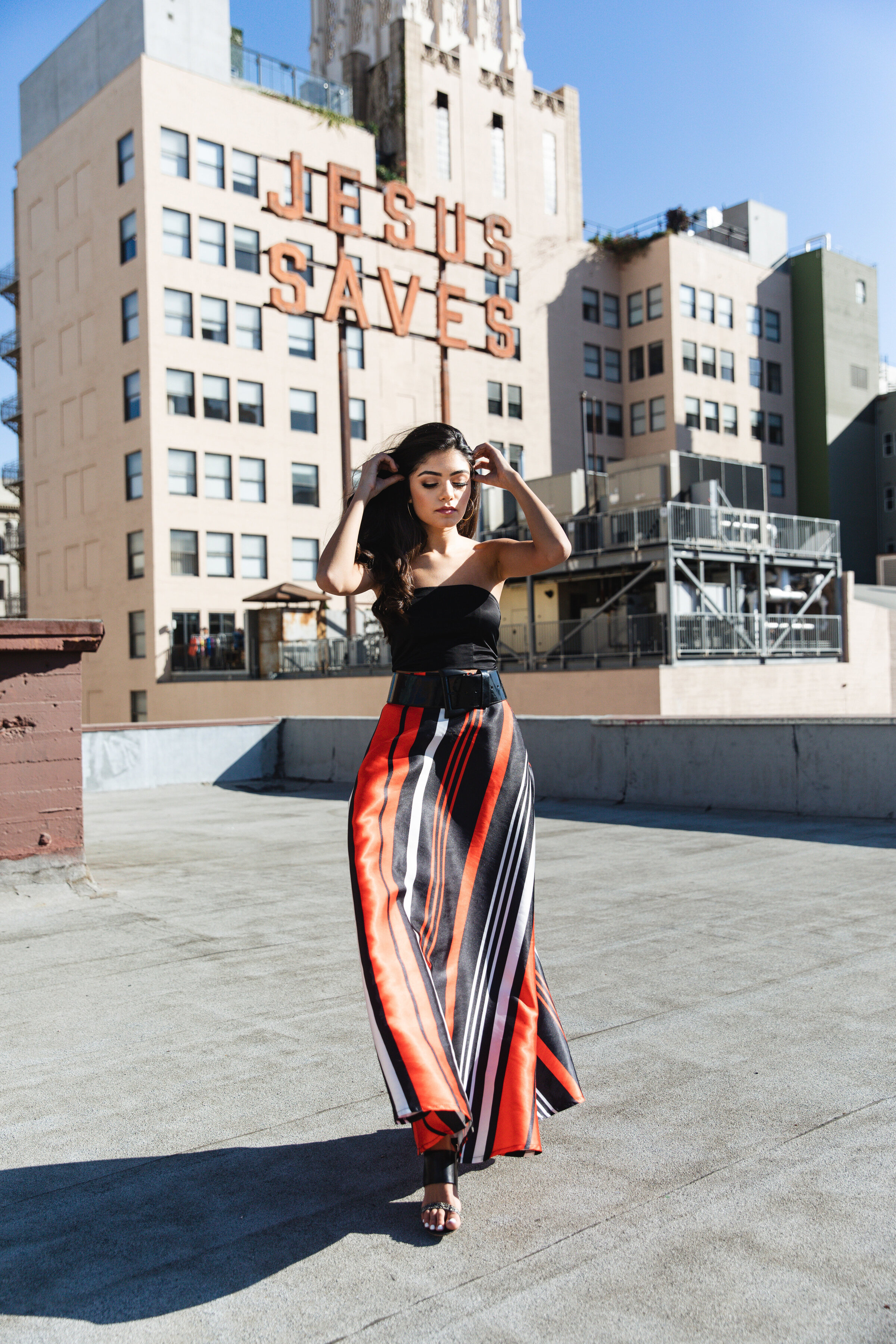 Rooftop editorial portrait of a woman in a strapless black top and bold red-and-black striped maxi skirt. She holds her hands in her hair with confidence, with an urban skyline and a vintage neon “JESUS SAVES” sign in the background.
