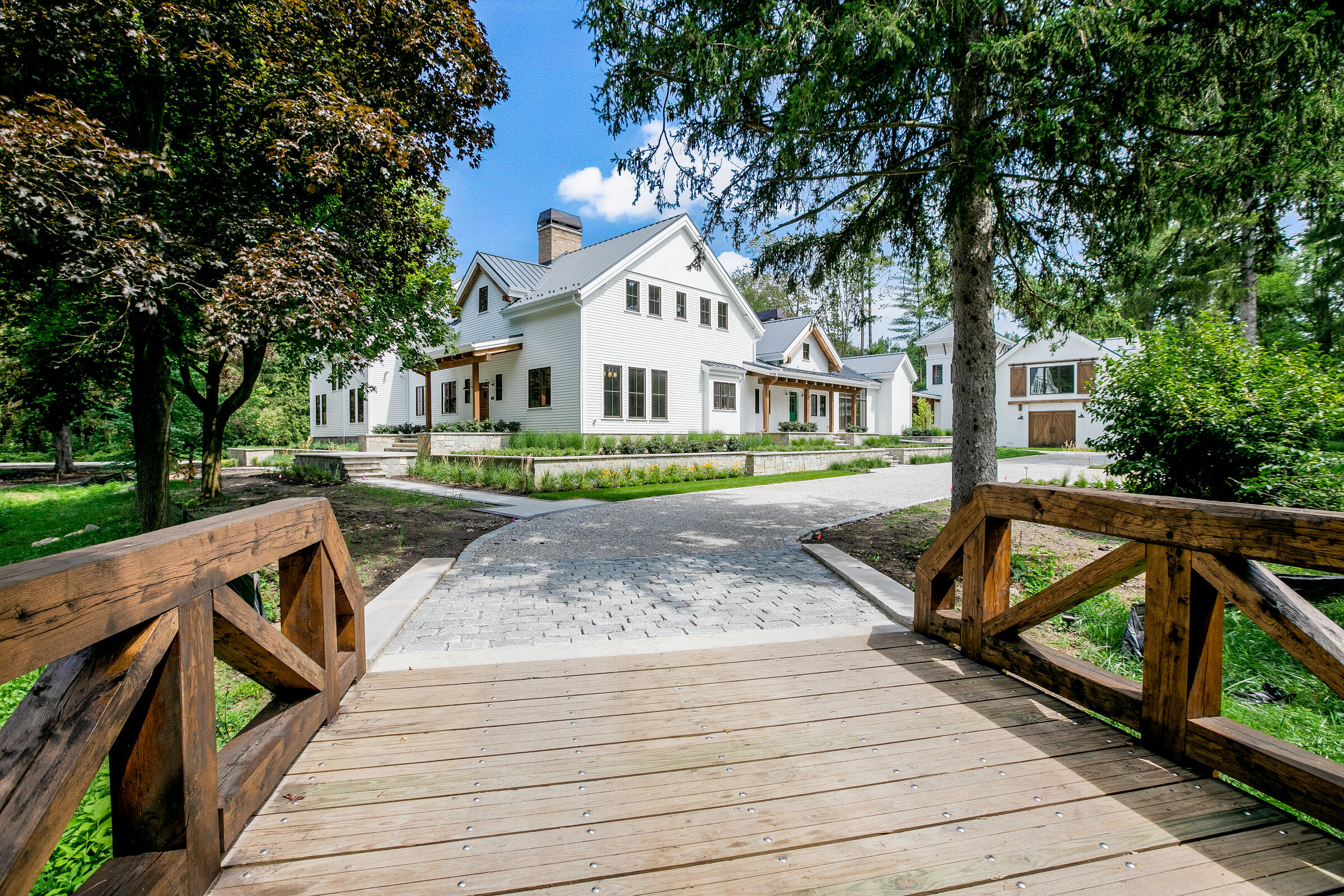 A breathtaking view from a bridge overlooking a massive custom-built home surrounded by tall trees and lush green grass. The long paved driveway leads to the stately residence, showcasing the grandeur and natural beauty of the property.