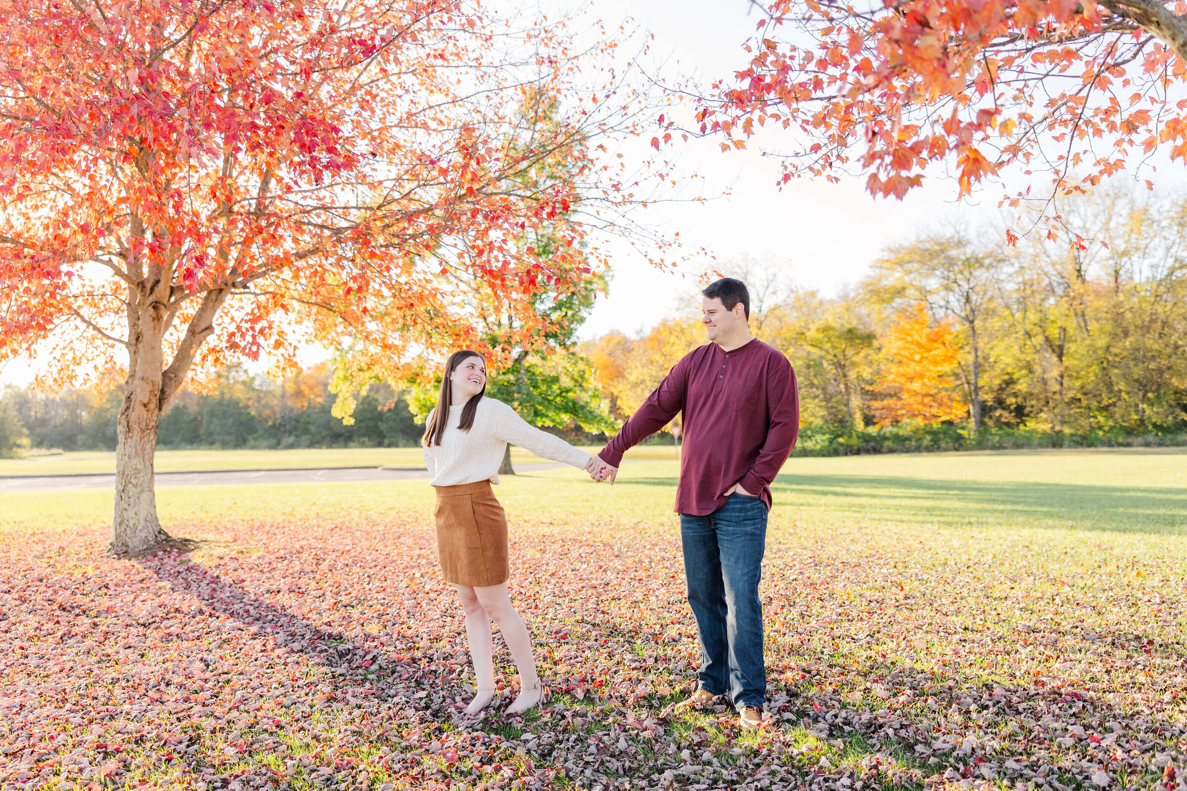 A woman holds a man's hand as they walk in a park. The trees are bright red because it's fall in Ohio. She is wearing a tan skirt with an ivory blouse and he is wearing blue jeans and a maroon shirt.