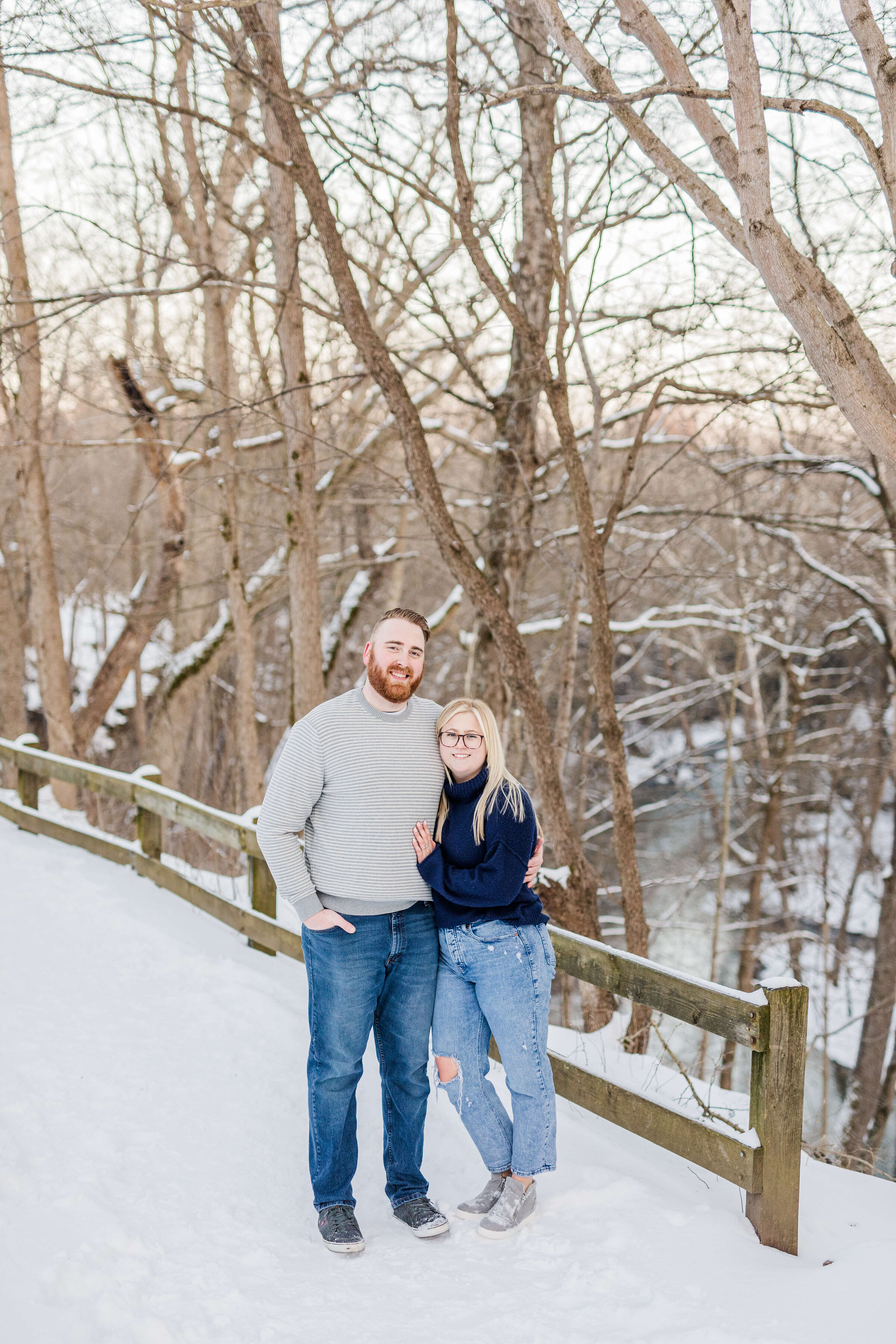 A man and woman stand next to each other with the man's arm around the woman. They are standing in the snow surrounded by trees and there is a river below them