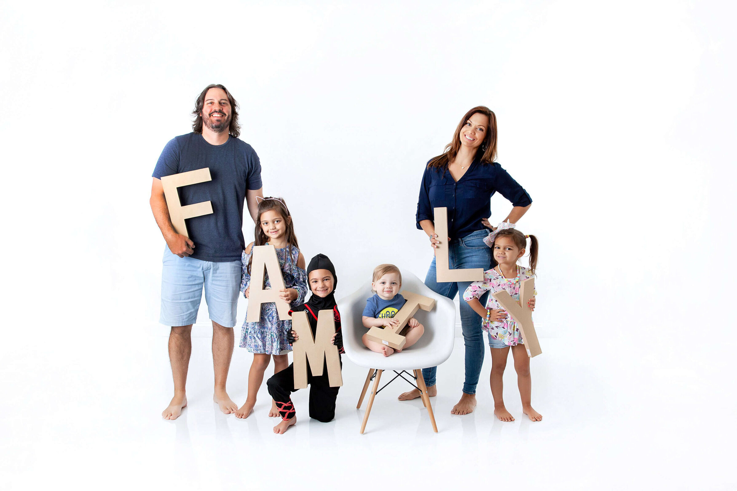 A family of six poses against a white background at a Jacksonville portrait studio. The parents stand on either side, holding the letters "F" and "Y." Four children hold the letters "A," "M," "I," and "L." The child holding the "O" sits on a chair. They are all smiling and barefoot, capturing a joyful moment with their Jacksonville family photographer.