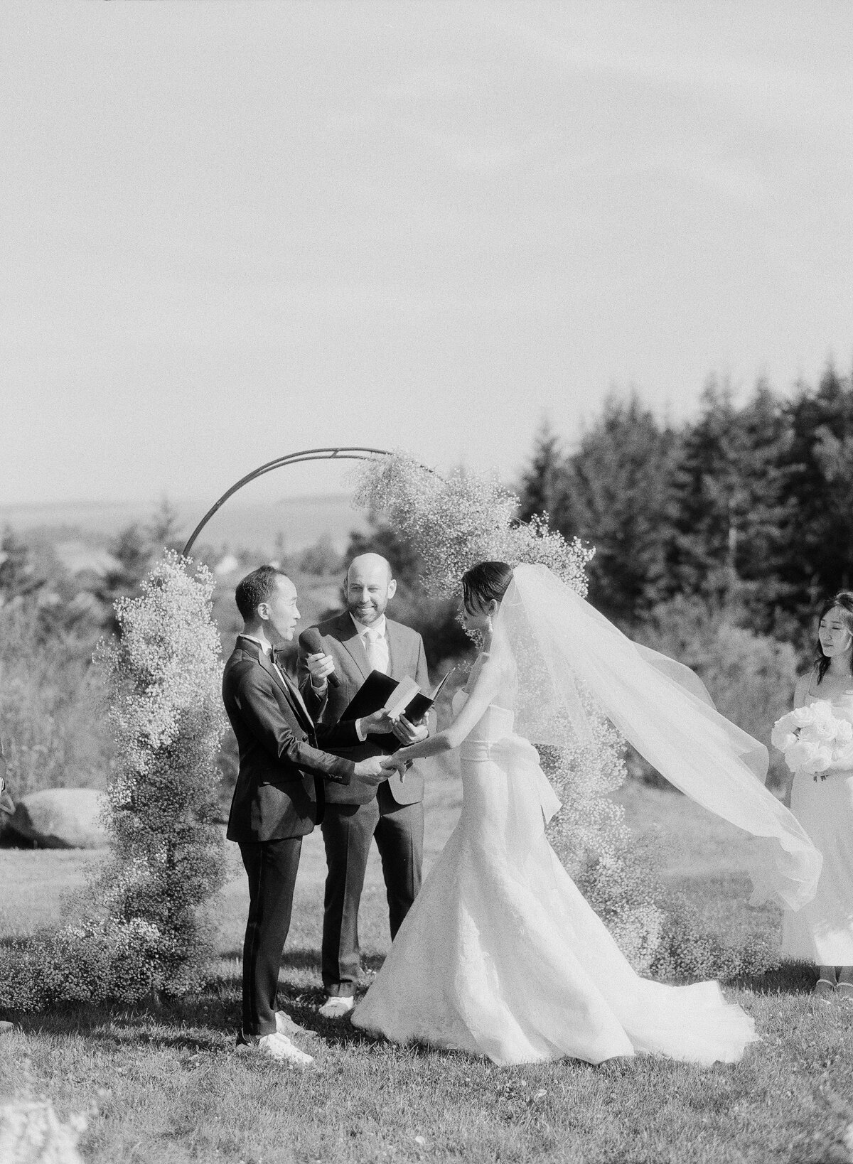 black and white photo of bride and groom at the ceremony outdoors with veil blowing in the wind