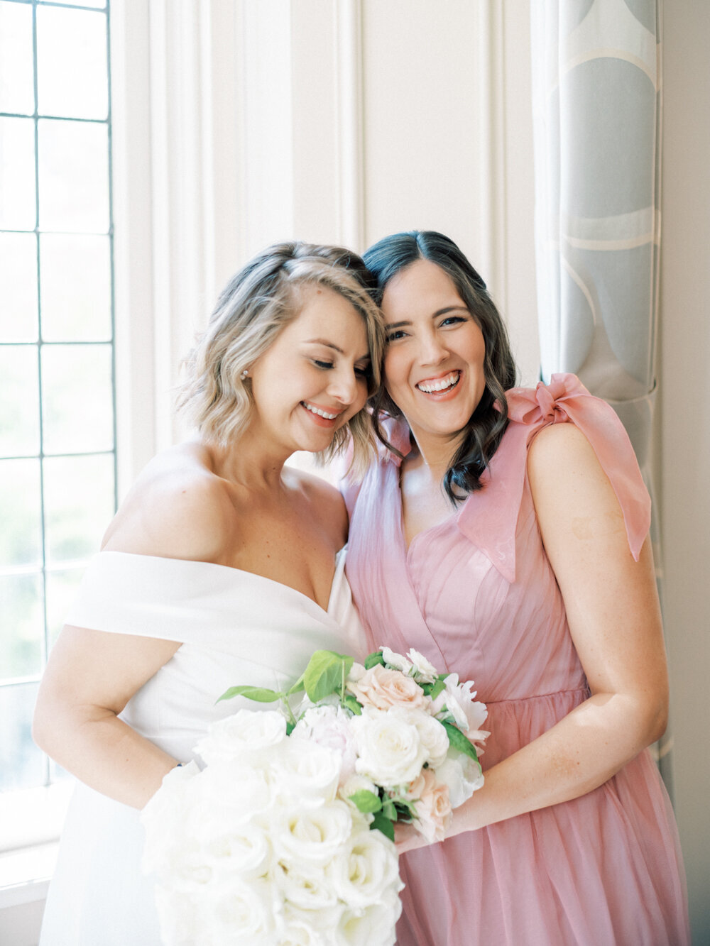 bride with off the shoulder white gown hugging her bridesmaid in a pink chiffon gown