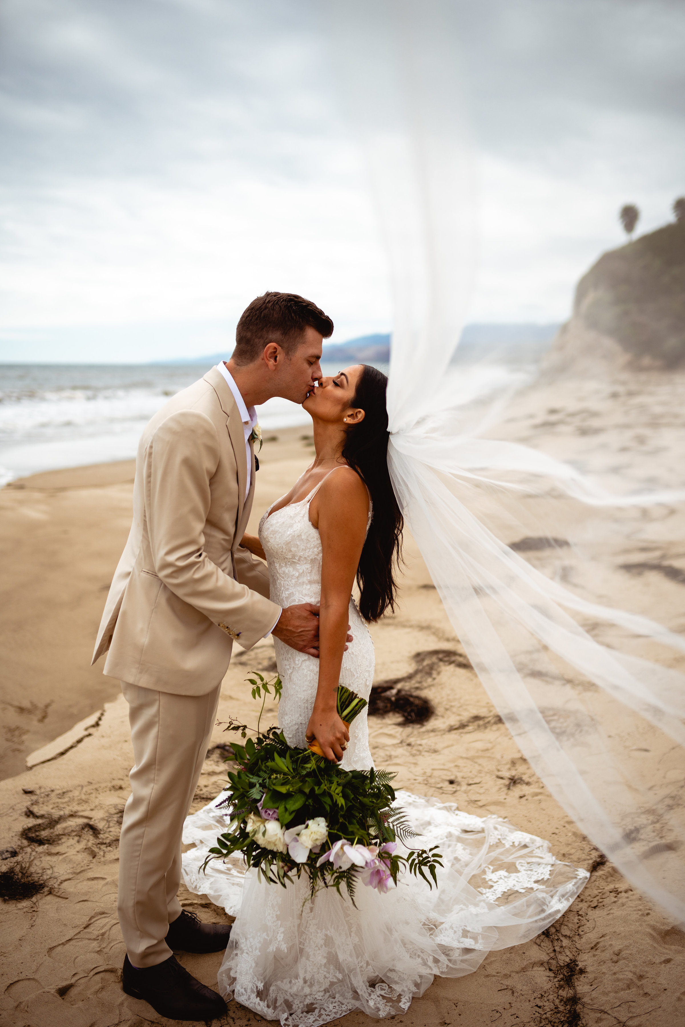 groom kisses the bride with veil blowing in wind on beach