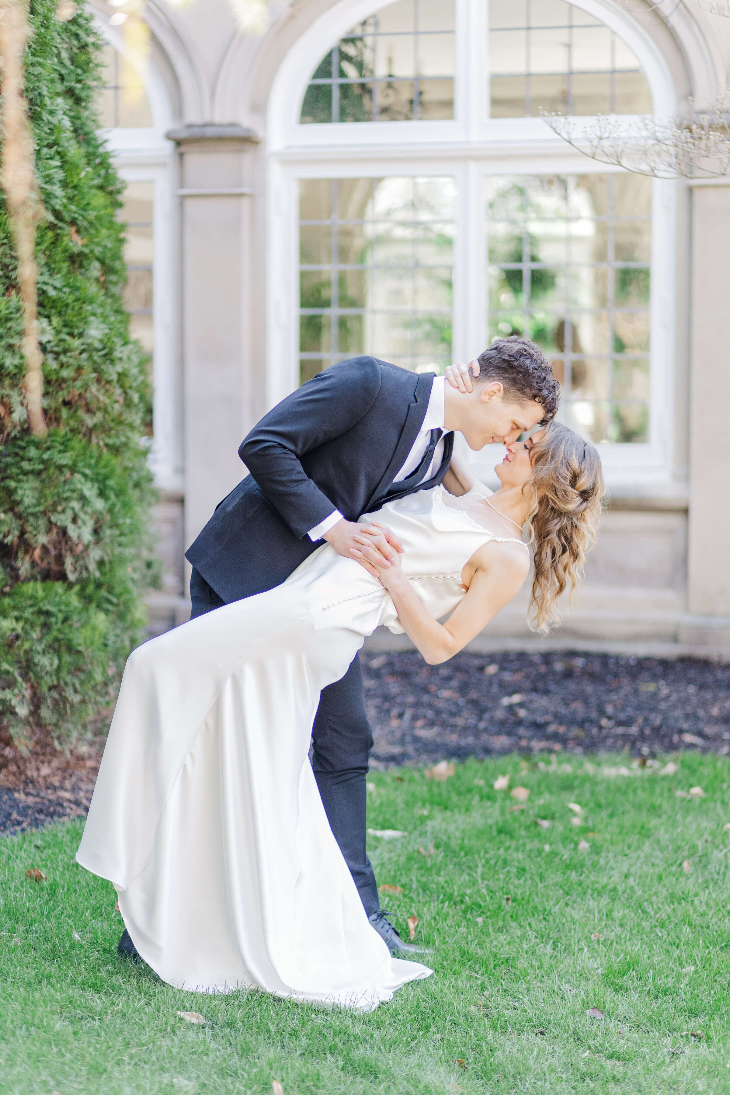 A groom in a black suit dips his bride back as they look at each other. They are outside on the green grass in front of a mansion.