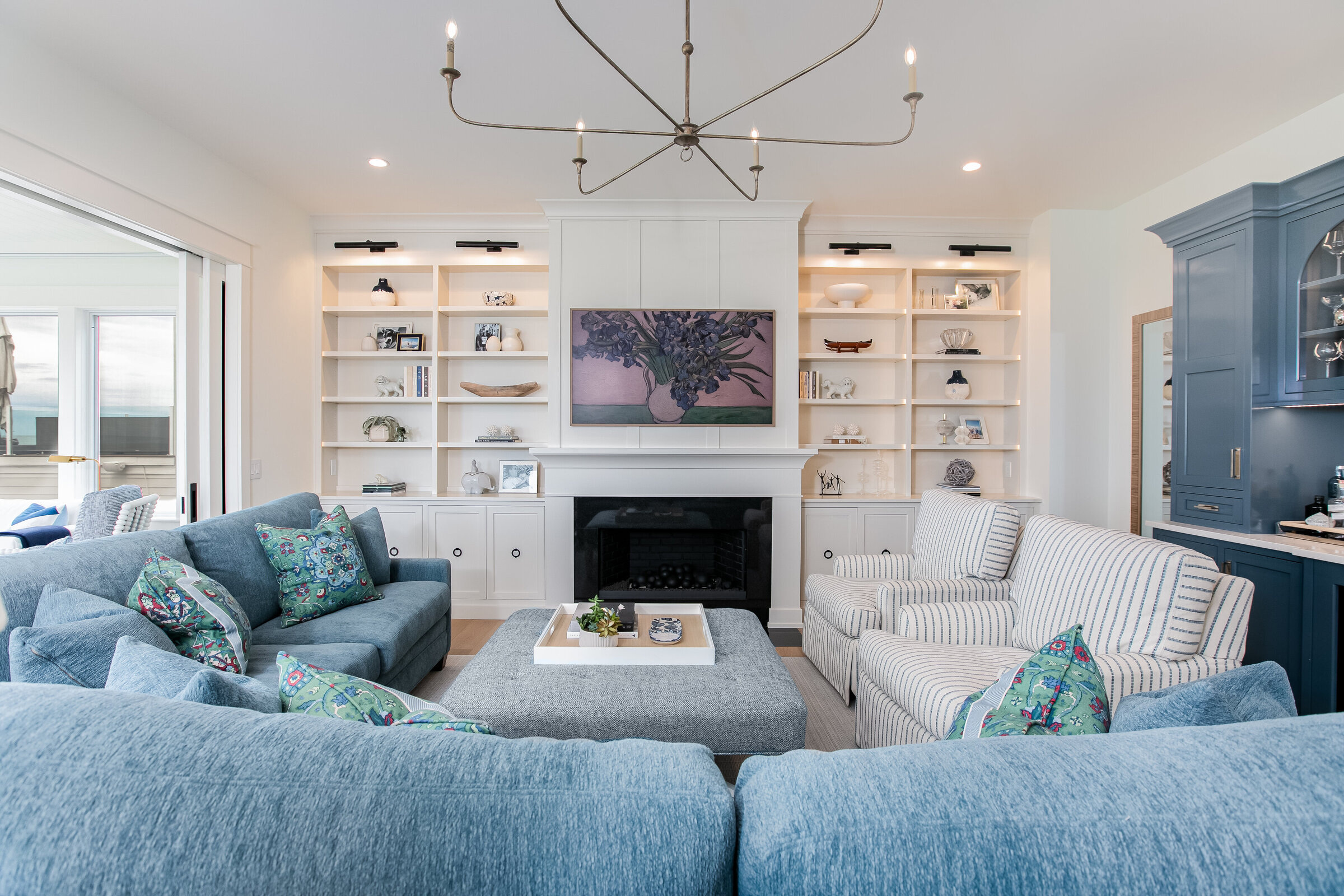 A beautifully designed living room featuring open shelving built-ins on each side, illuminated by overhead lights. The space is anchored by a simple yet massive chandelier and a blue couch adorned with decorative cushions, creating a cozy and stylish environment.