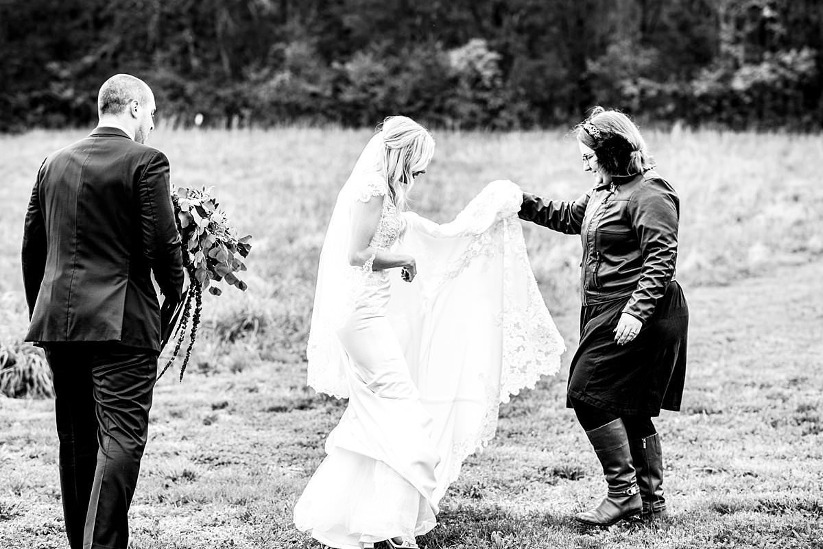 Black and White photo of Mahlia helping bride walk in her wedding dress through field at Drakewood Farms
