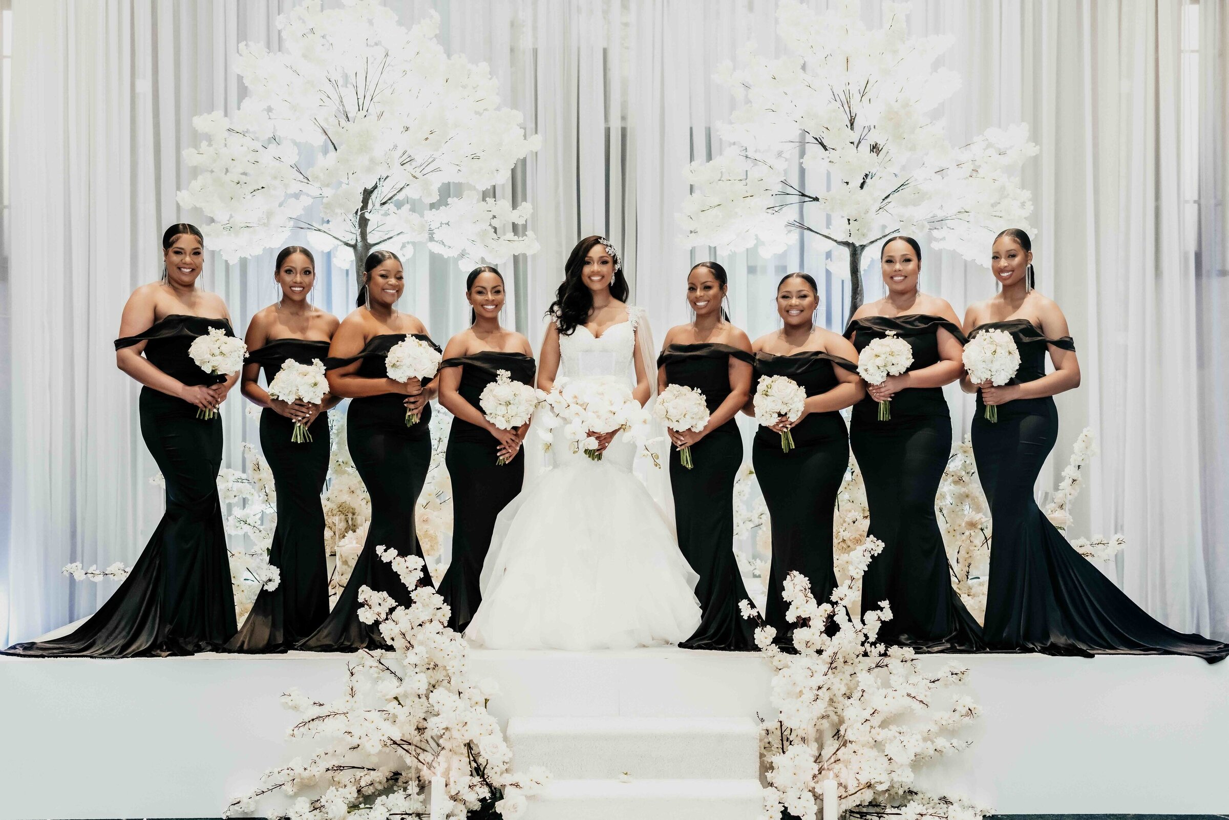 Bride stands with bridesmaids in black satin dresses holding white bouquets, framed by white floral trees and soft draping at a JW Marriott Nashville wedding.