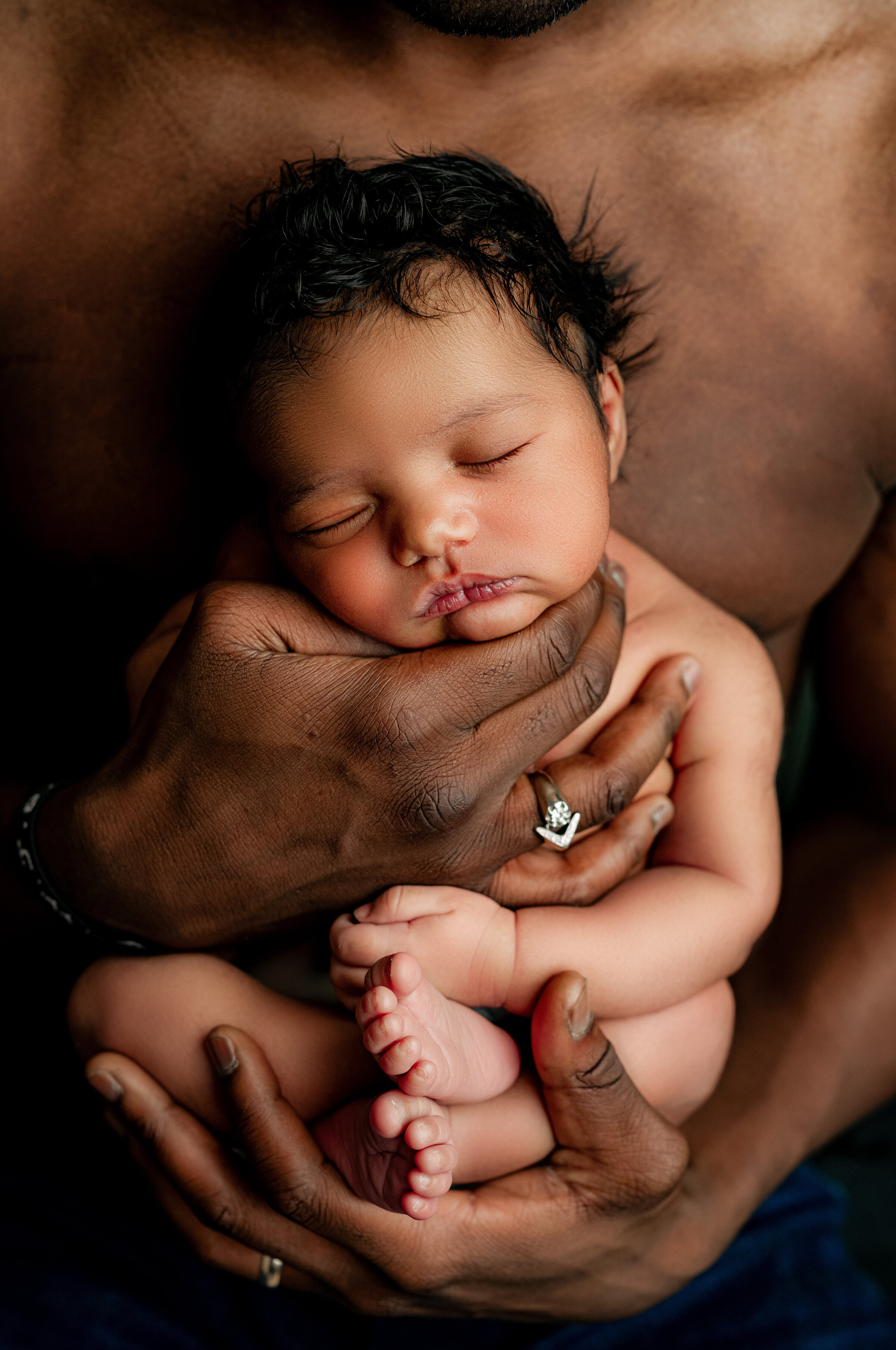 A newborn baby sleeps peacefully, cradled in the arms of an adult with dark skin. The babys tiny hands and feet are visible, while the adults hands provide a gentle and protective embrace. This Charlotte newborn photography scene conveys warmth and tenderness.