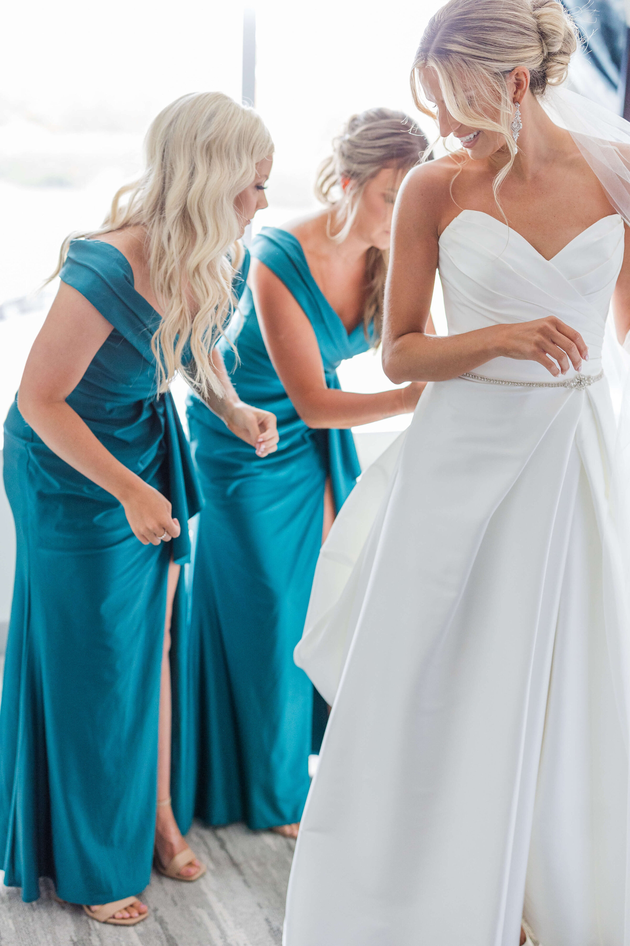 A bride stands with her bridesmaids as they help her with her dress. She's smiling and looking back at them. They are in teal dresses.