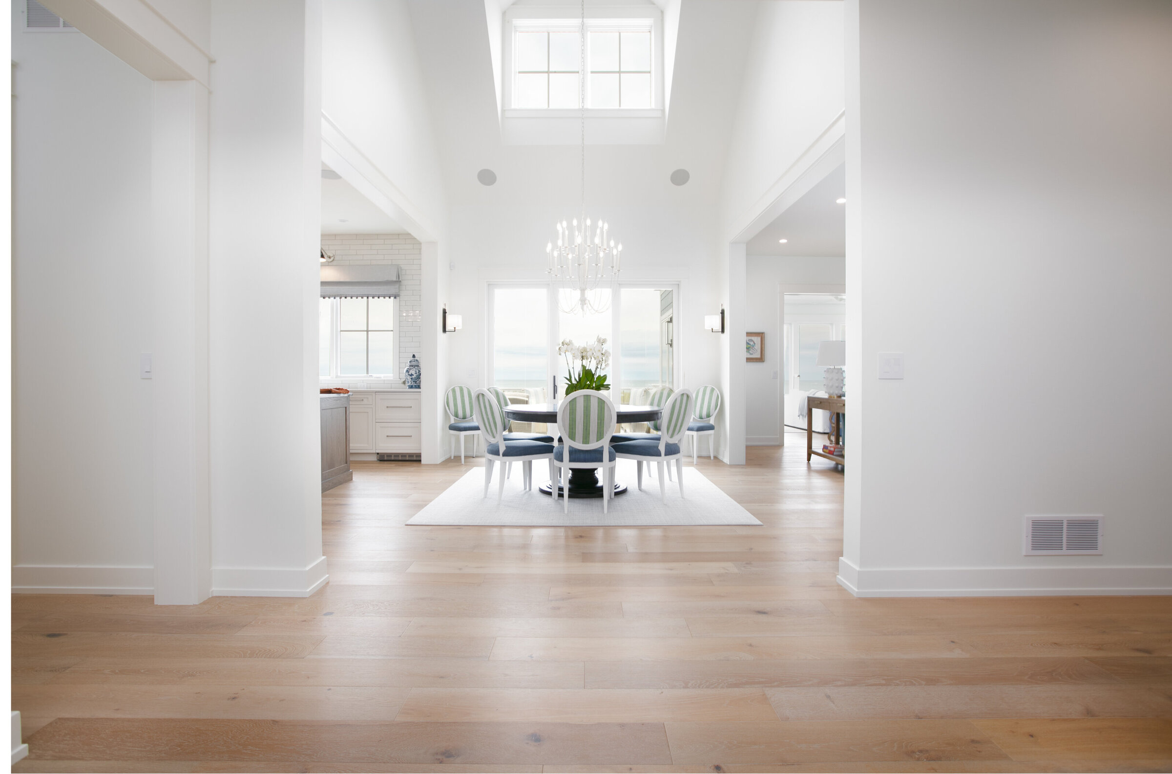 A majestic entryway featuring 30-foot ceilings, white walls, and skylights, offering a glimpse into a well-lit dining room with a round table and bold chairs. The design exudes elegance and openness, creating a stunning first impression.