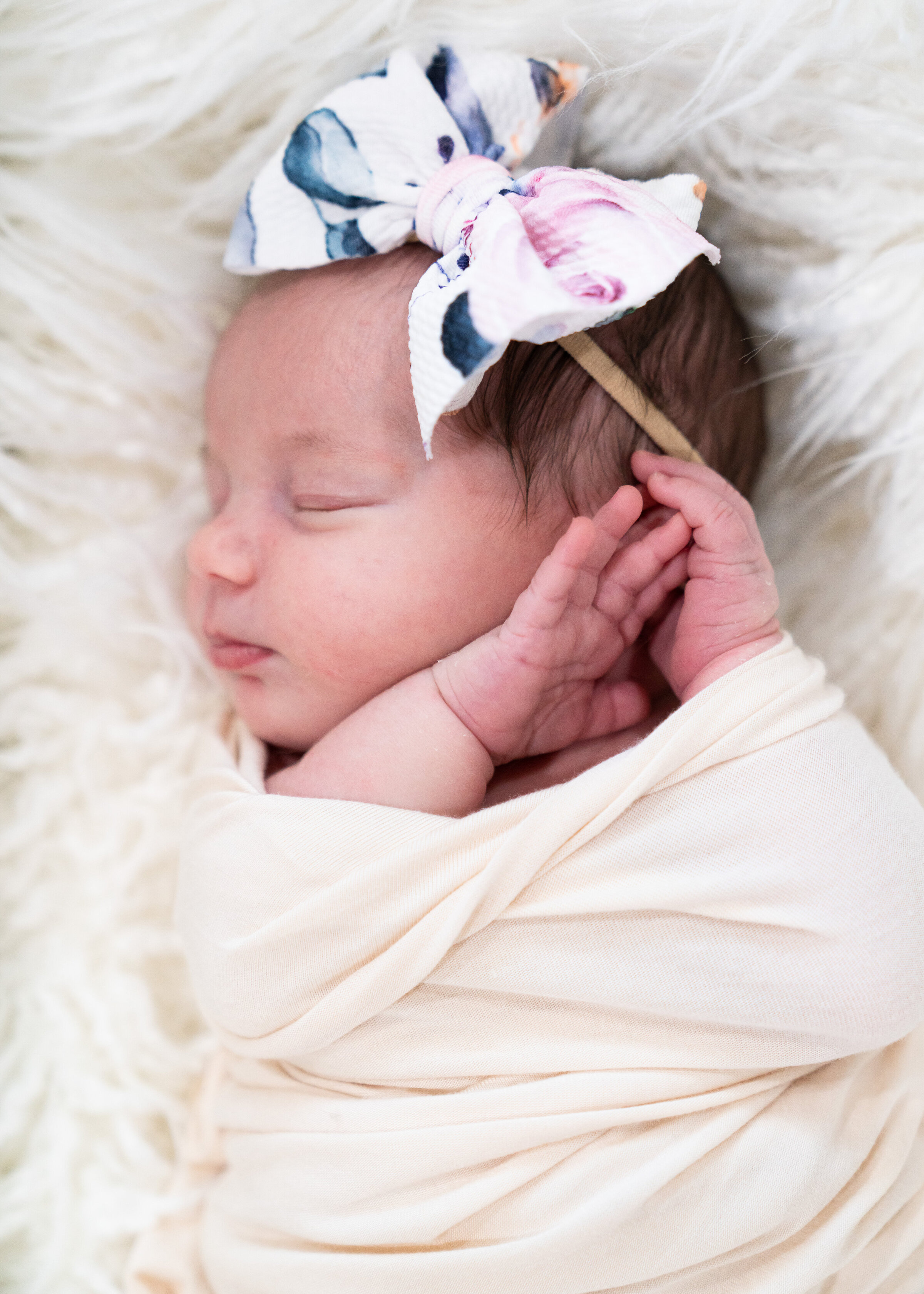 Newborn baby girl sleeping wearing a bow and wrapped in a cream colored cloth for her newborn photo shoot