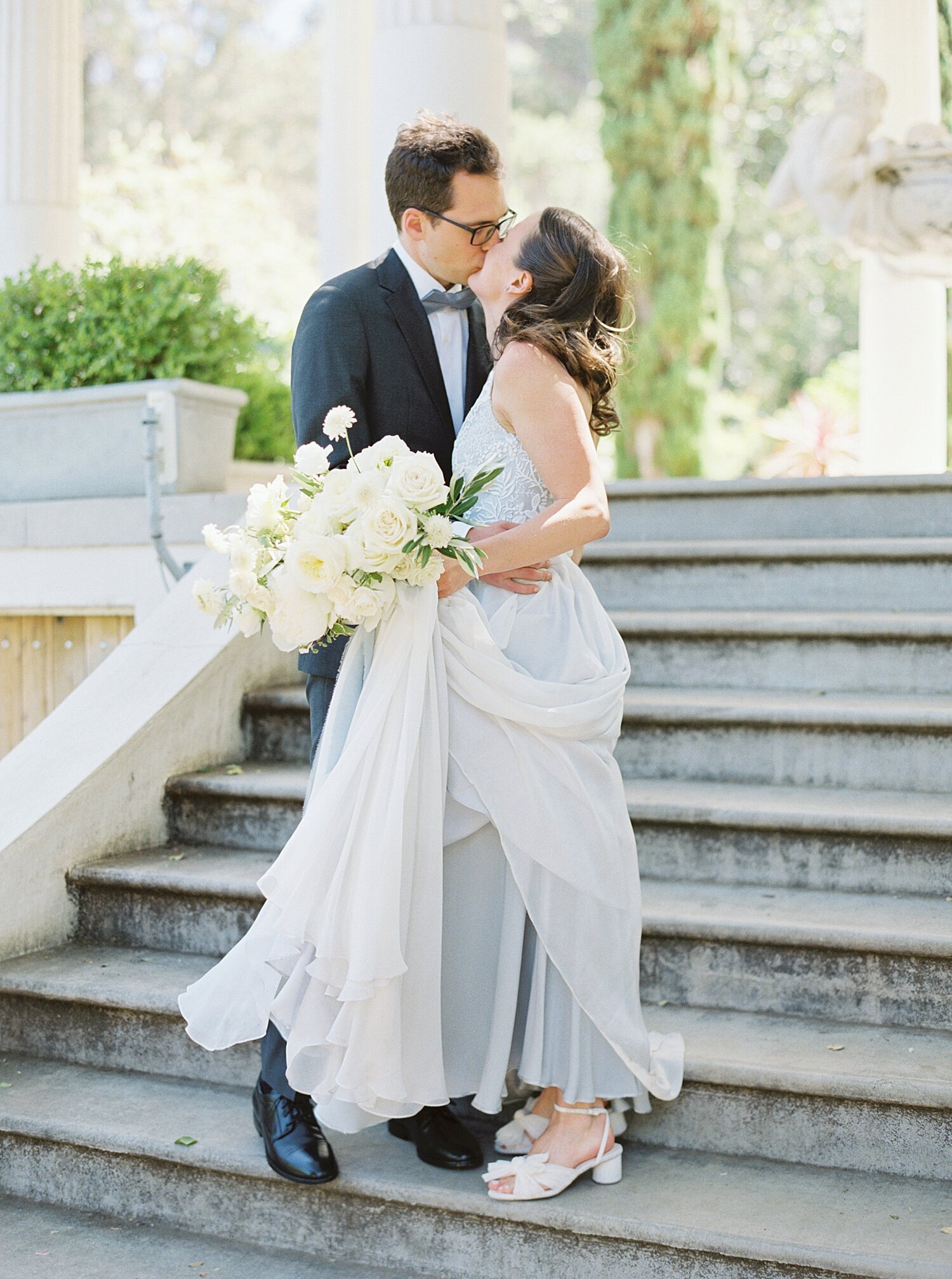 groom in navy suit kissing bride in light blue dress while holding white roses on the stairs