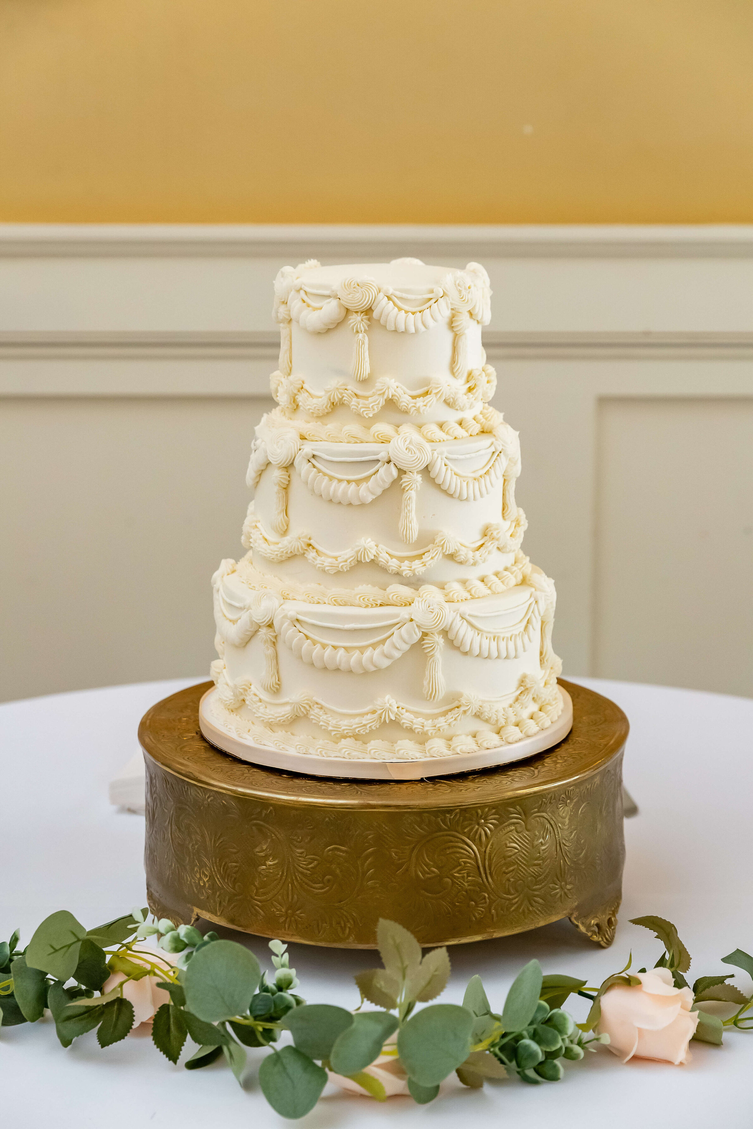 A three-tiered white wedding cake with ornate icing decorations sits on a gold cake stand, placed on a white table with green eucalyptus leaves and a pale pink rose.