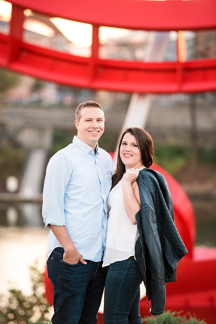 Engaged couple smiling at camera with the artistic red sculpture Tumbleweed and the Cumberland river behind them