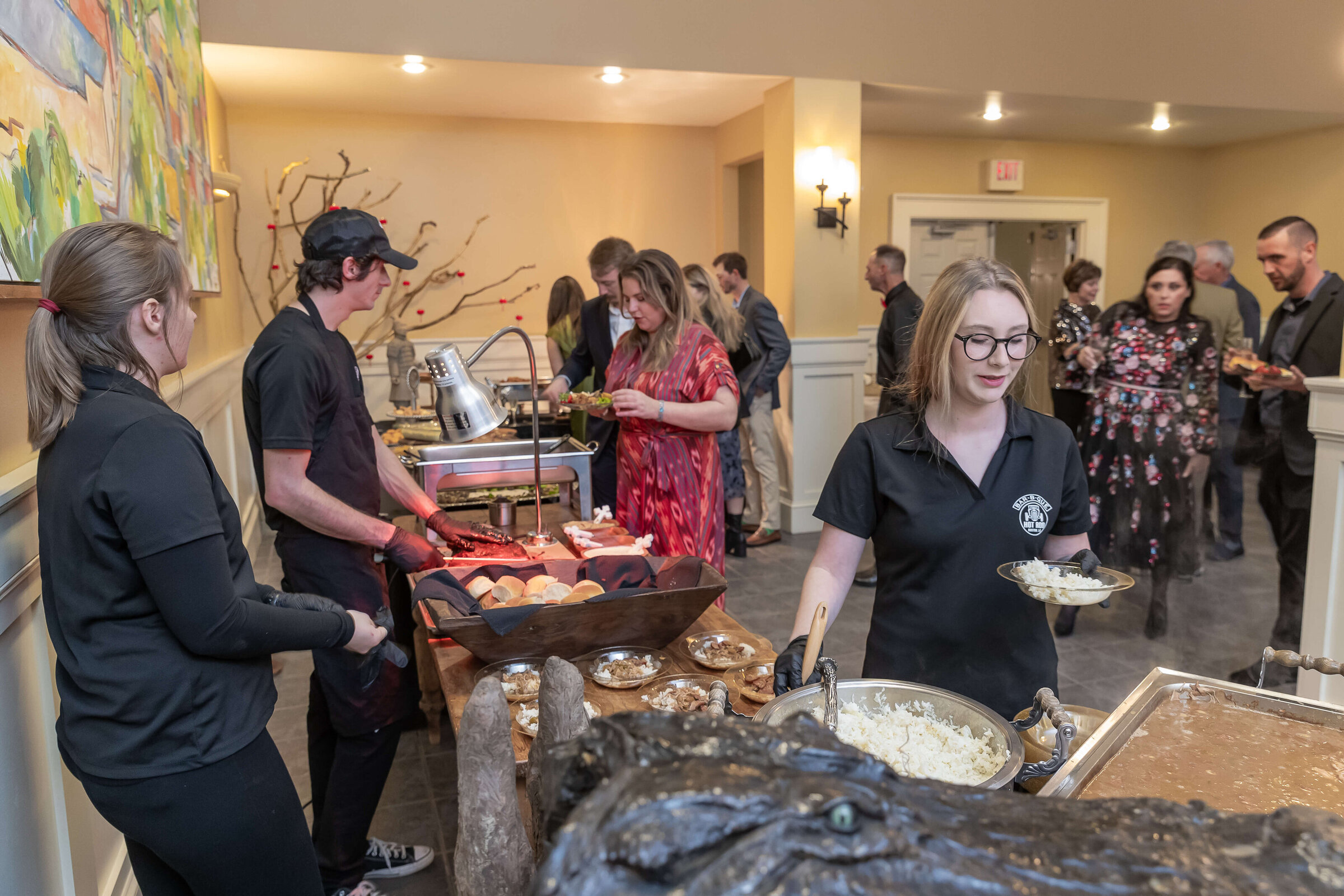 People at a buffet in a warmly lit room; servers in black uniforms assist as guests help themselves to food. Some guests are chatting near the buffet table, while others are selecting dishes.