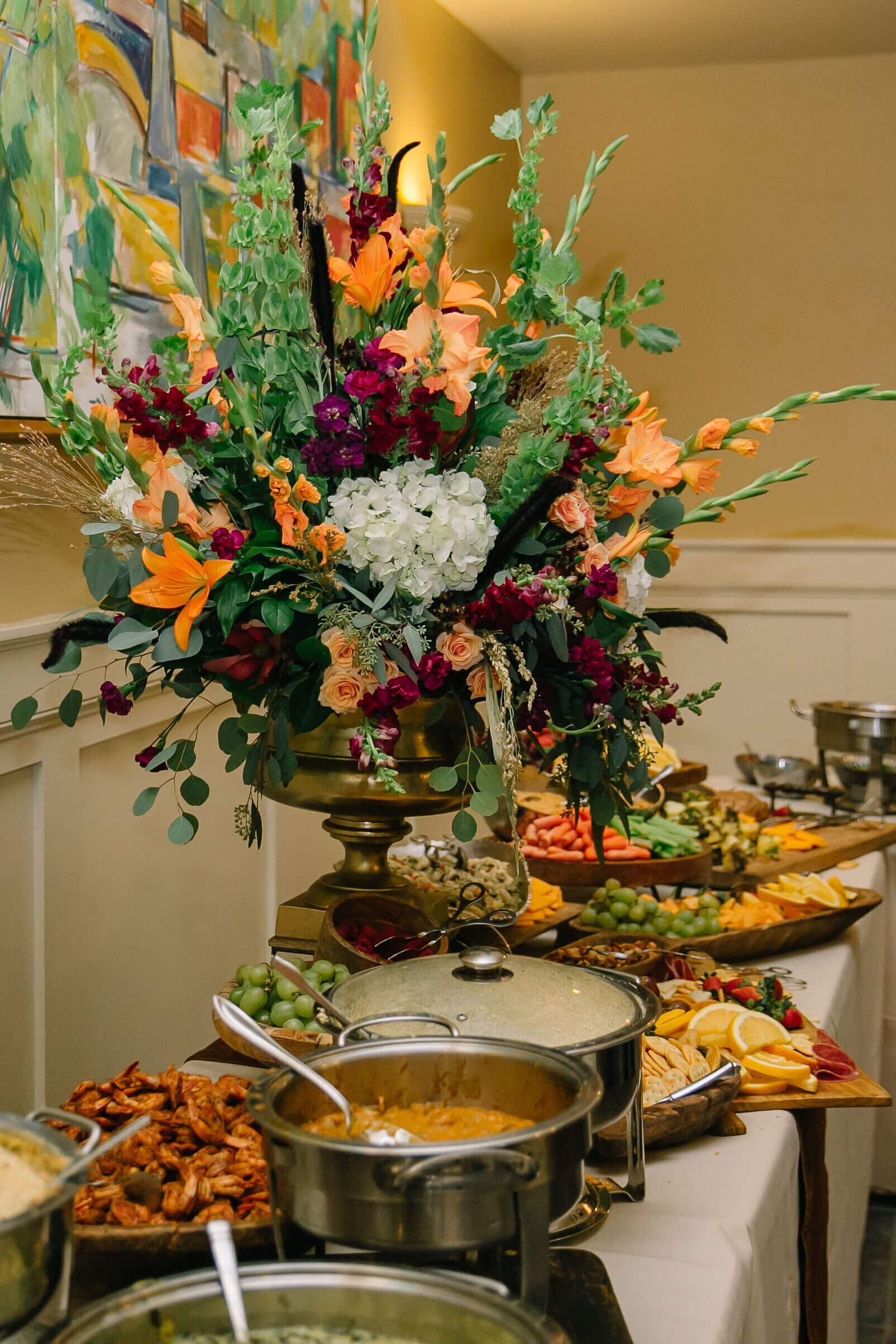 A buffet table with a wooden tiered stand holding assorted fresh fruits and leafy floral arrangements, surrounded by cheese, crackers, vegetables, and a tray of sandwiches in a bright, elegant room.