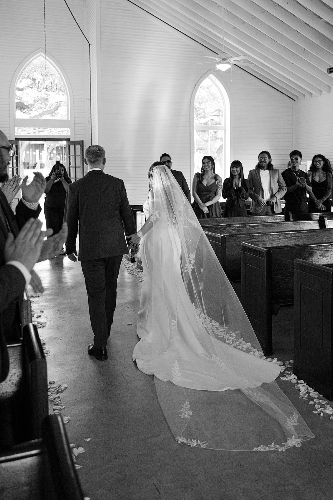 Bride escorted down the aisle in a classic Texas chapel at The Chandelier of Gruene as guests rise, captured in timeless black and white.