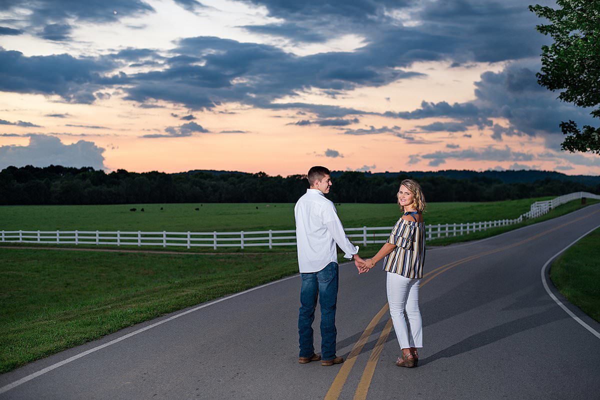 Couple strolling down a country road with white fences and a cloudy sunset rolling in the distance