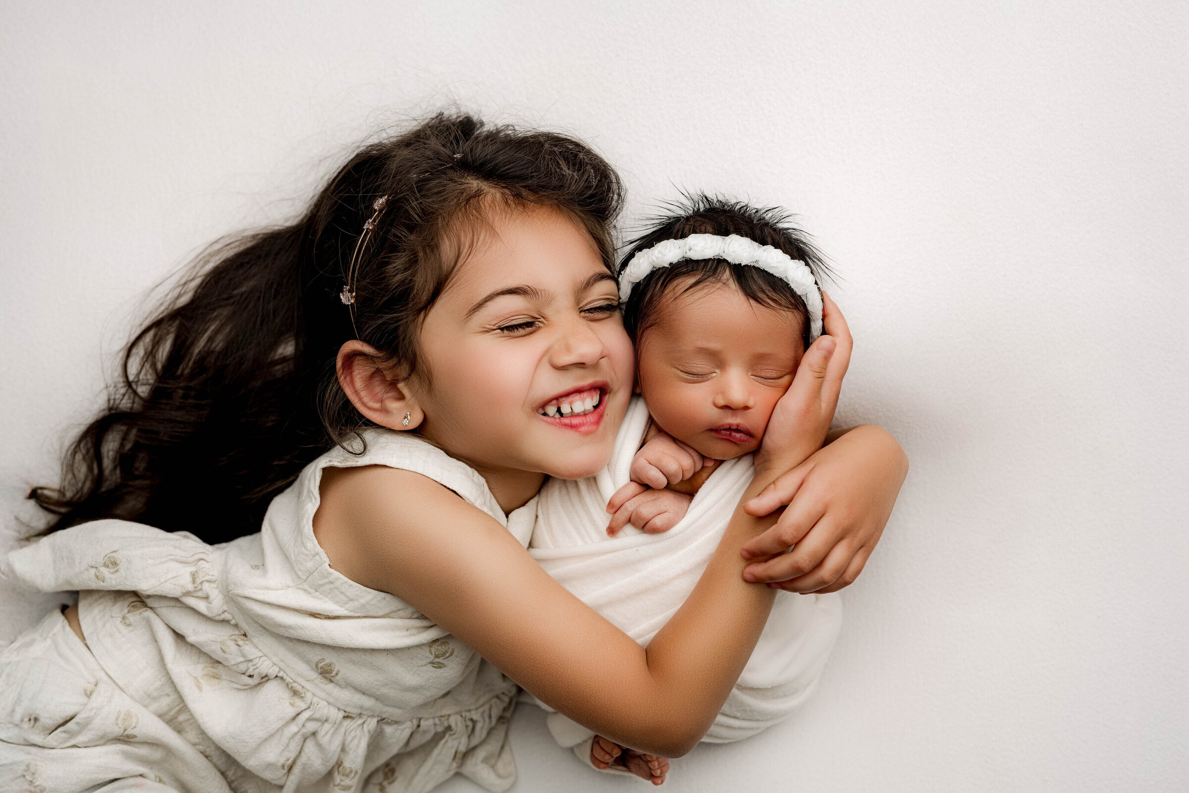 Big sister joyfully hugging her swaddled newborn sibling, both smiling and cozy on a white background.