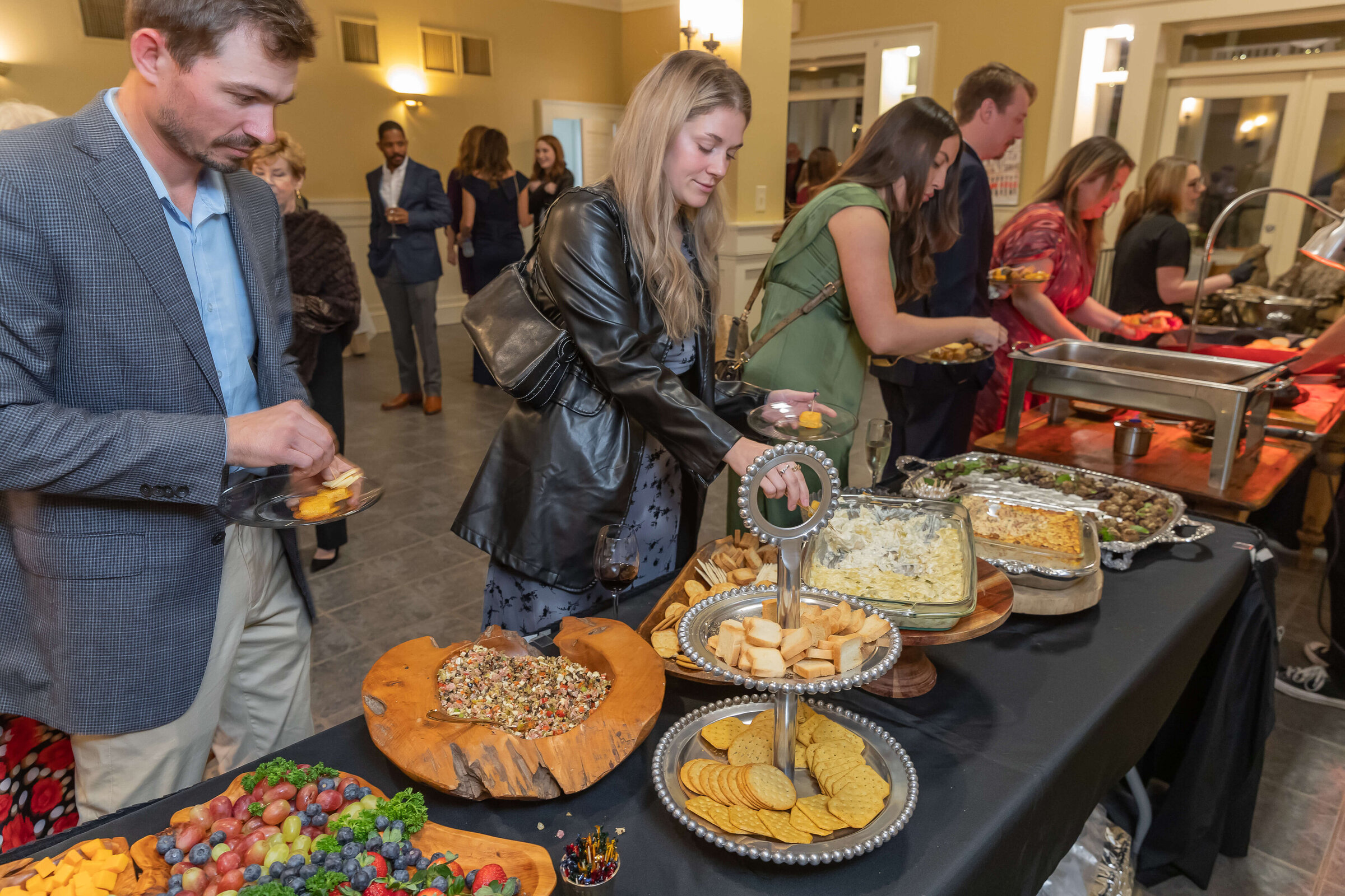 People dressed in semi-formal attire serve themselves food from a buffet table with a variety of dishes, including fruit, crackers, dip, and hot entrees, at an indoor social event.