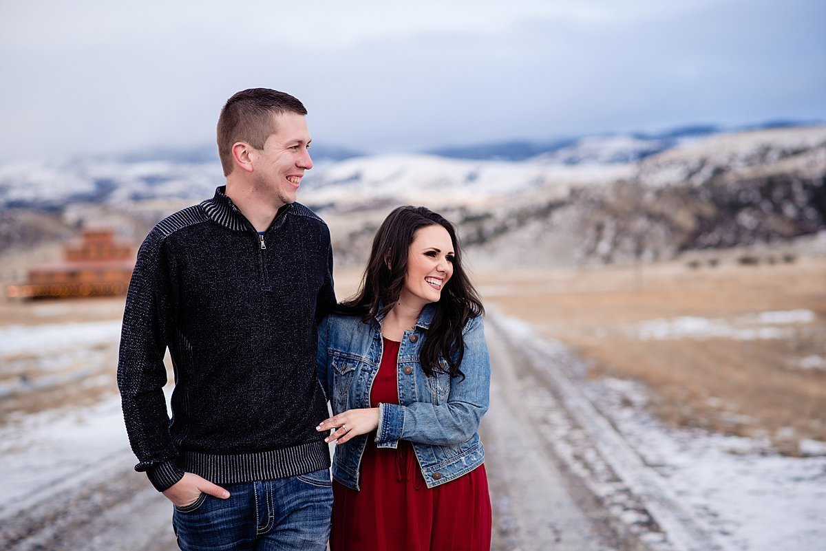 Close up photo of engaged couple laughing and smiling off into the distance with snow coming down and mountains behind them near Bozeman Montana