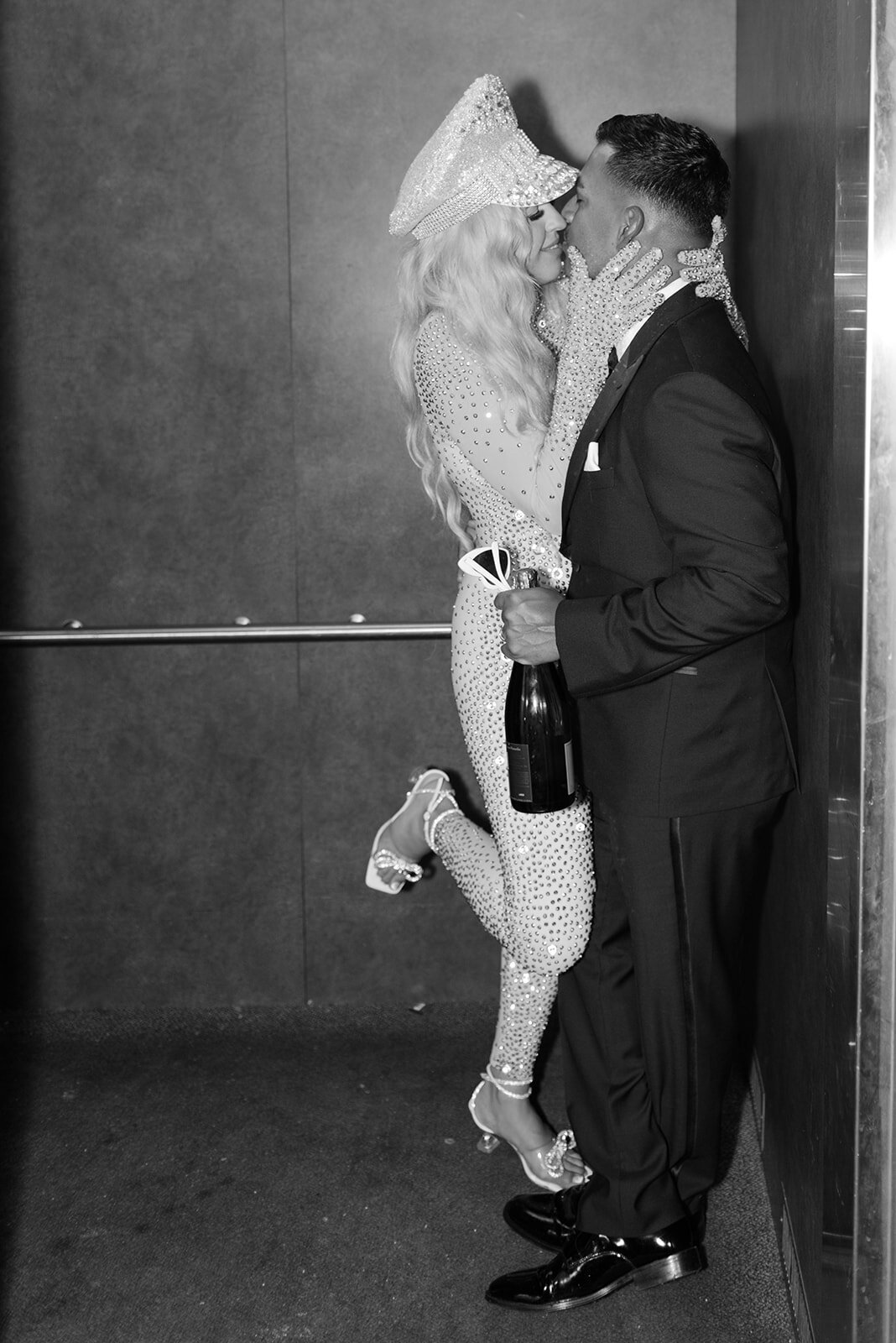 Editorial black and white portrait of bride and groom in an elevator, sharing a kiss after their Austin wedding reception while holding a champagne bottle in hand