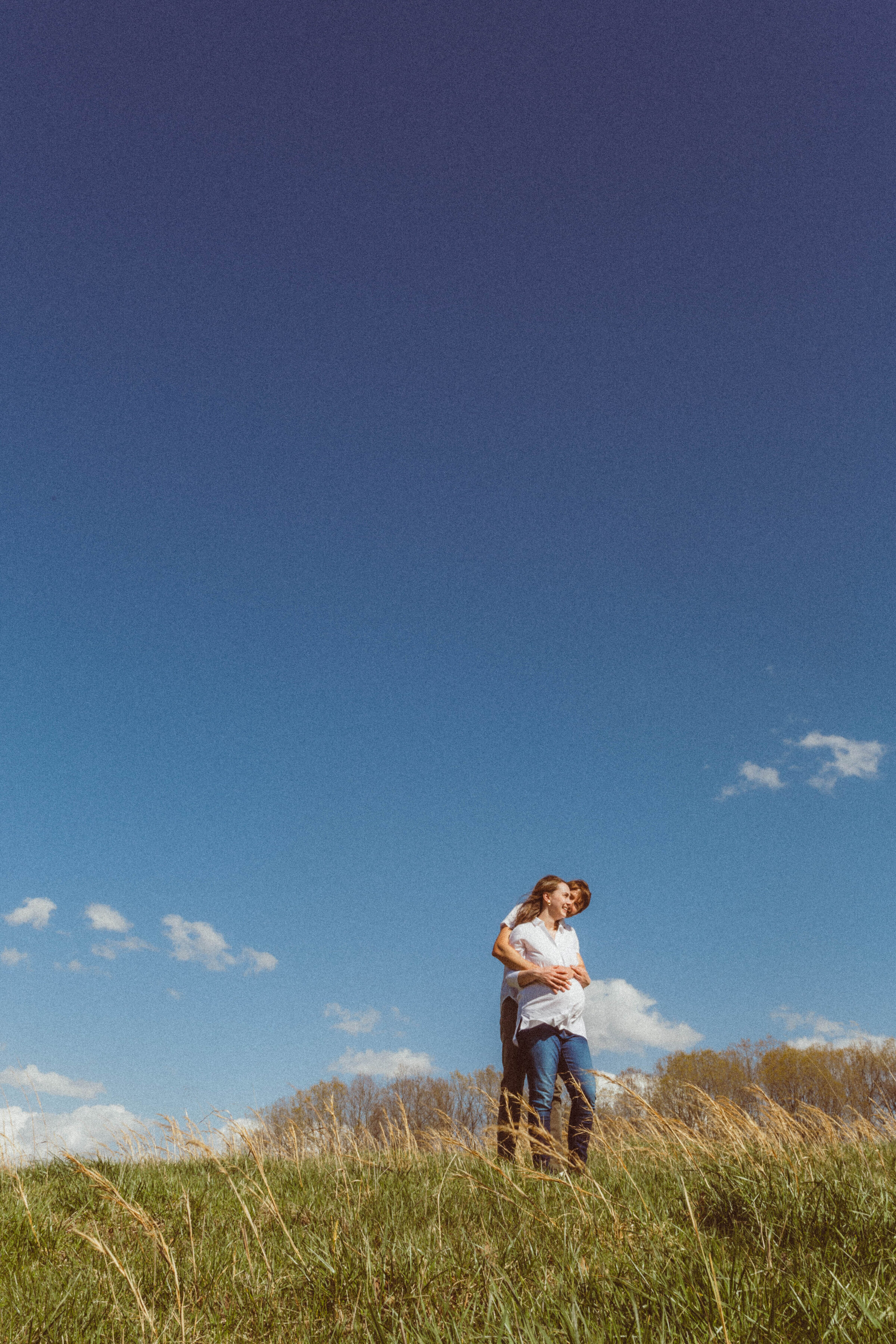 Far-off shot of a husband and pregnant wife hugging with almost all sky as the backdrop, captured in a destination maternity portrait style by Emily Ambie.