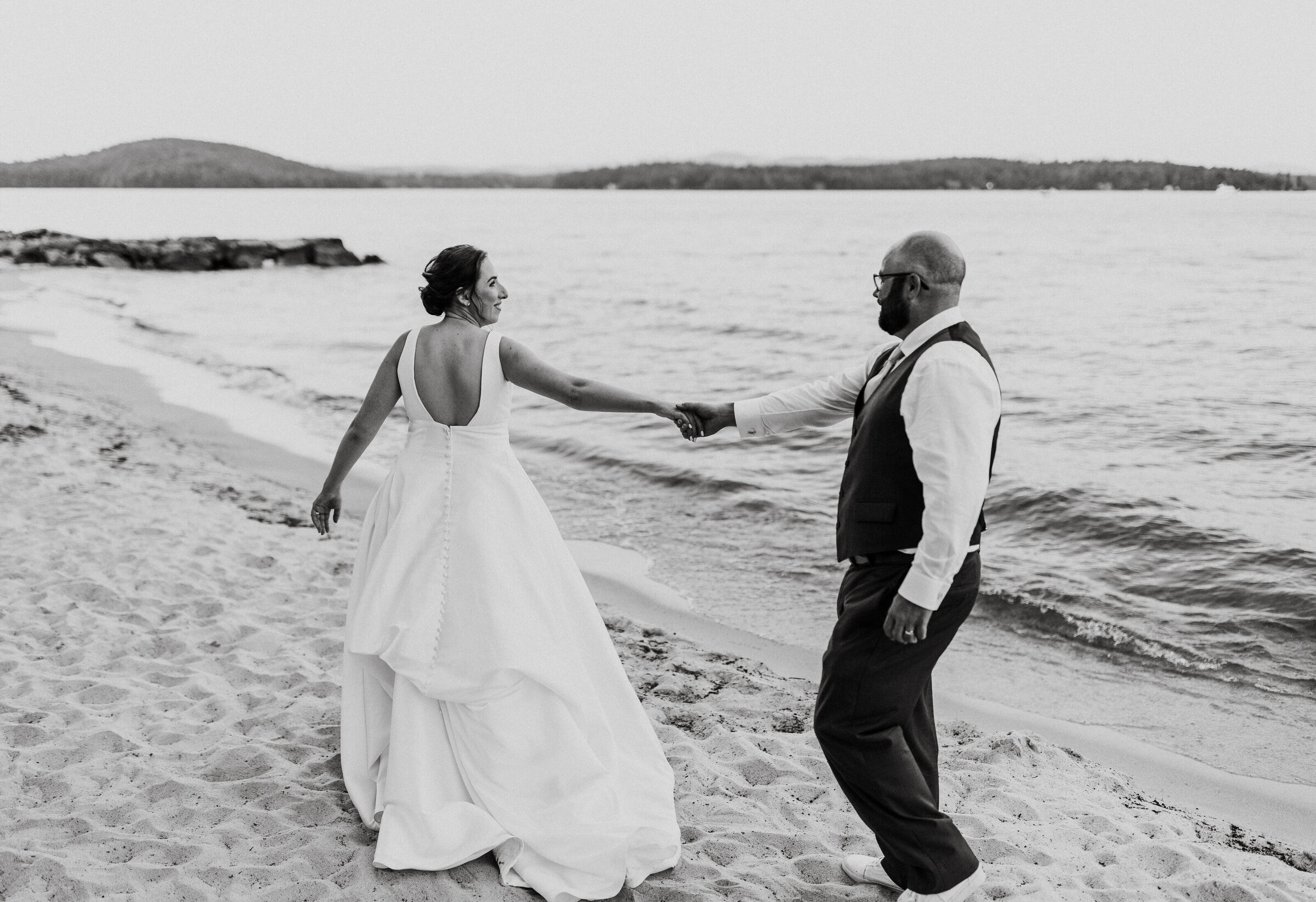 Bride and groom hold hands and walk along the beach at summer wedding in New Hampshire
