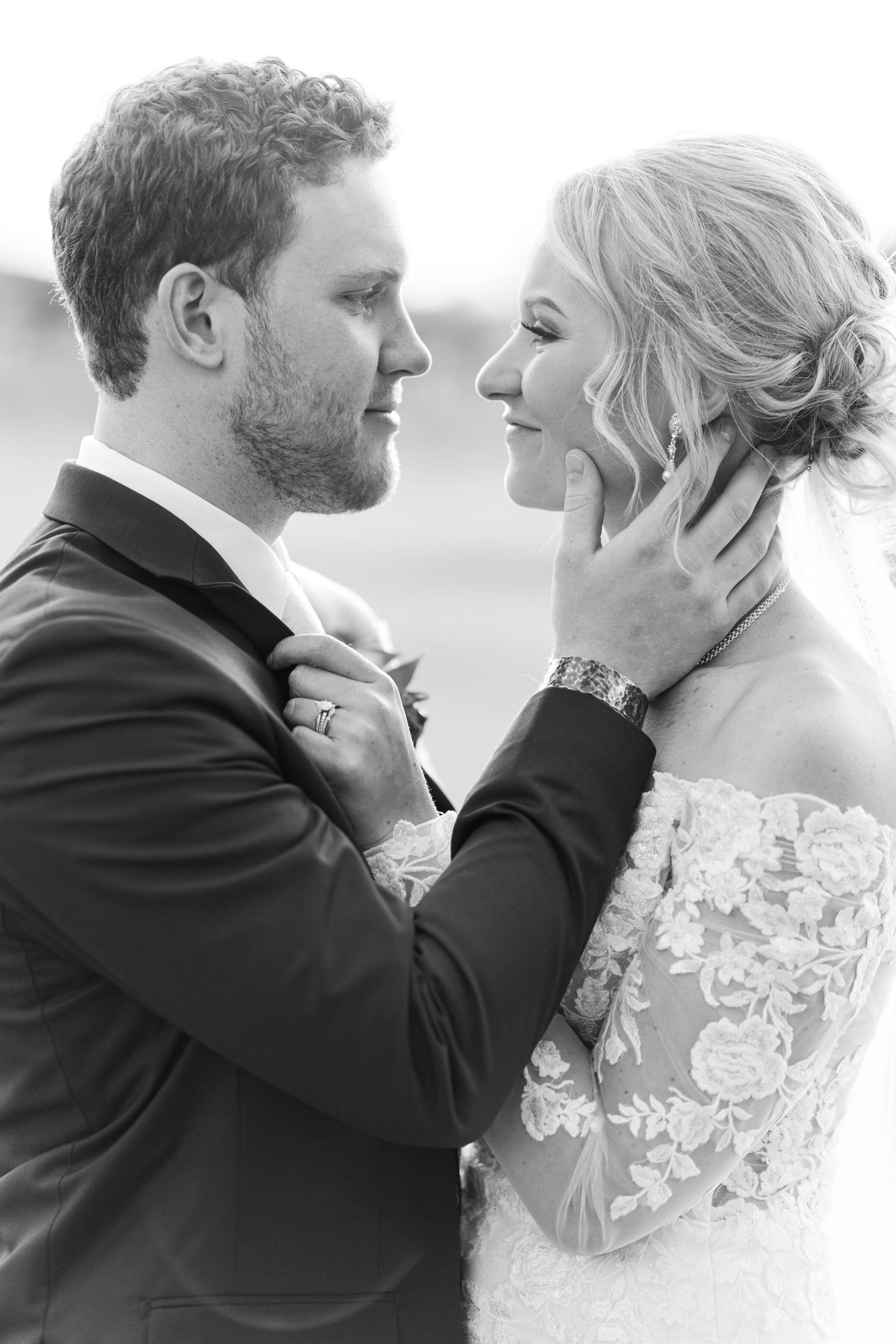 A black and white image of a groom and bride standing looking at each other. The groom has his hand on his bride's face as they come in for a kiss.