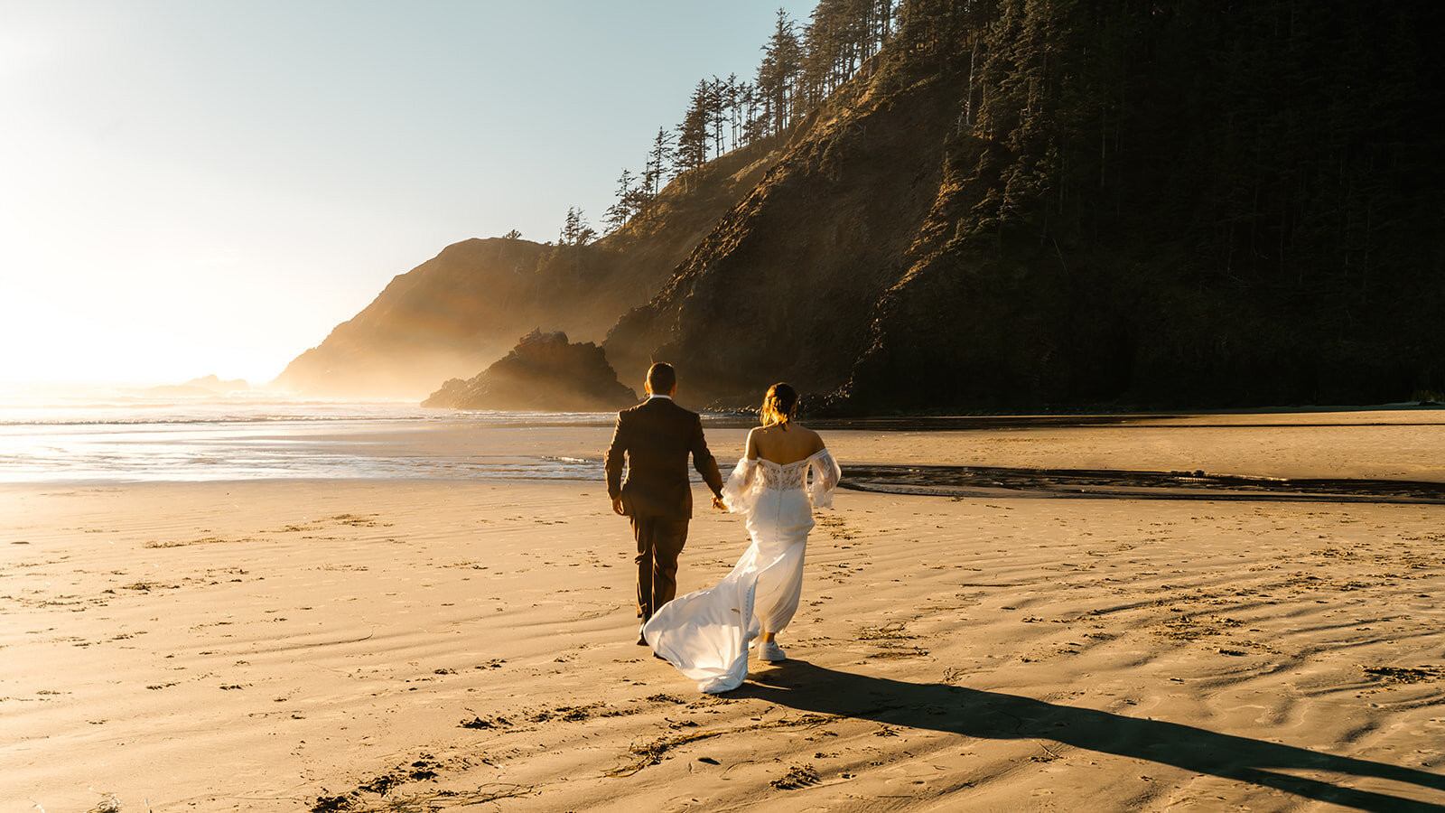 a couple in wedding attire walk away from their adventure elopement photographer and towards a rocky feature on the beach. They are painted in a golden light as the sun sets on the beach, the brides dress billowing behind her.