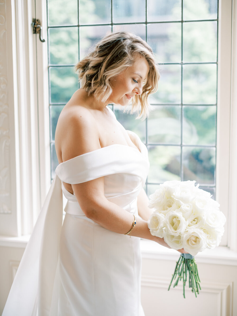portrait of bride in her off the shoulder gown with short blonde bob haircut and white long stem roses in front of large window