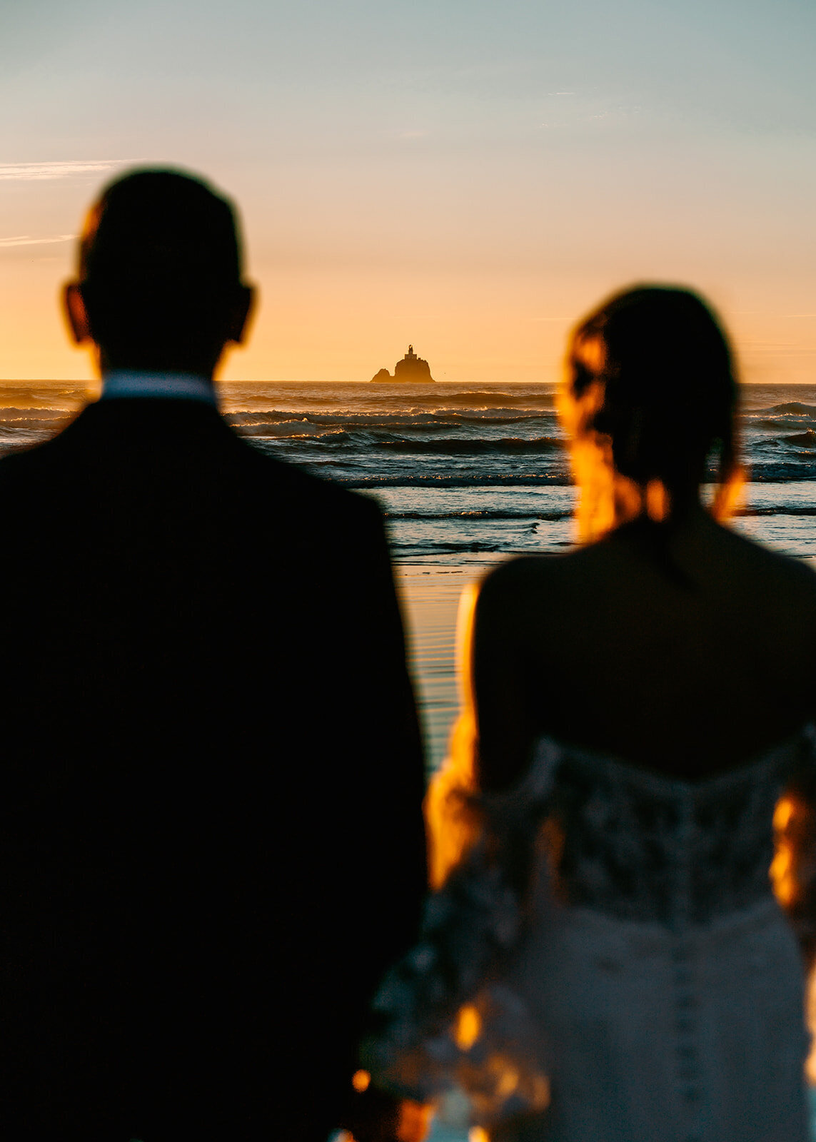 A couple, in their wedding attire gaze out at a lighthouse that appears suspended in the pacific ocean. The sunset paints everything in its path a pinky-golden hue, making the rolling waves look electric blue. Their adventure elopement photographers shoot between the couple, focusing on the lighthouse and using them to frame the lighthouse giving the perspective of seeing what the couple sees