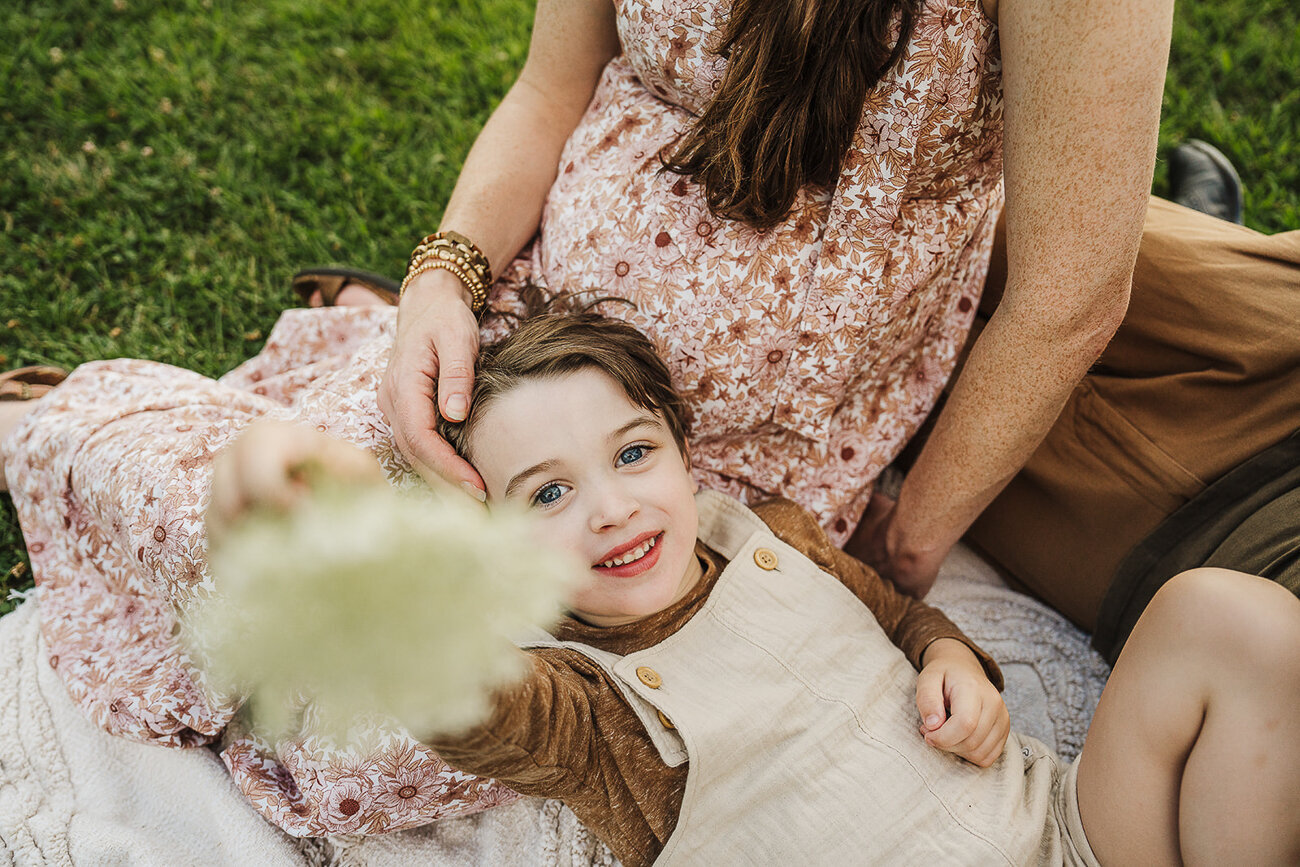 toddler brother lays on pregnant moms lap and smiles at camera