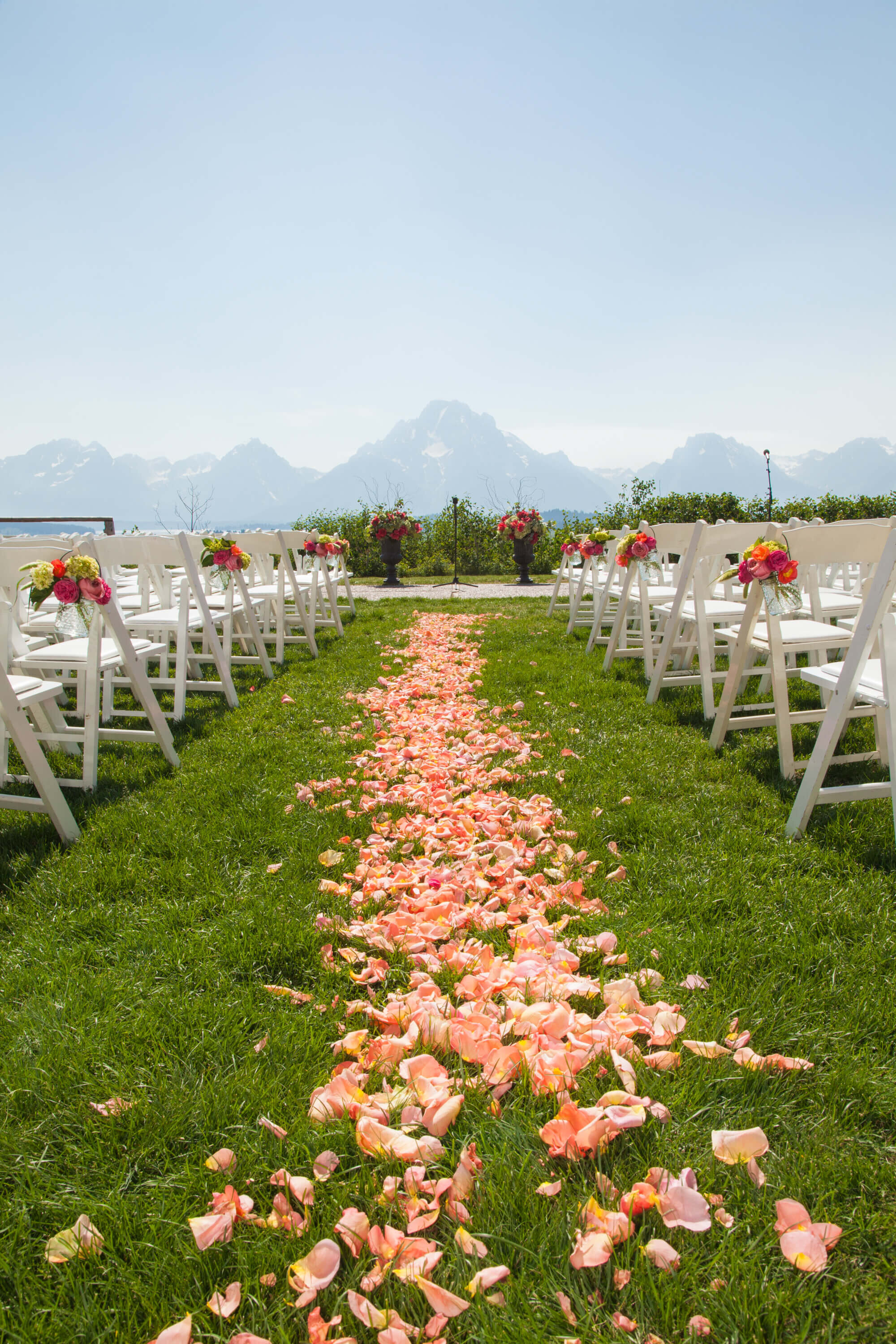 Outdoor Ceremony at Jackson Lake Lodge White chairs line a grassy aisle covered in rose petals, leading to a floral-adorned altar. The Grand Tetons provide a breathtaking backdrop for this Jackson Lake Lodge wedding ceremony.