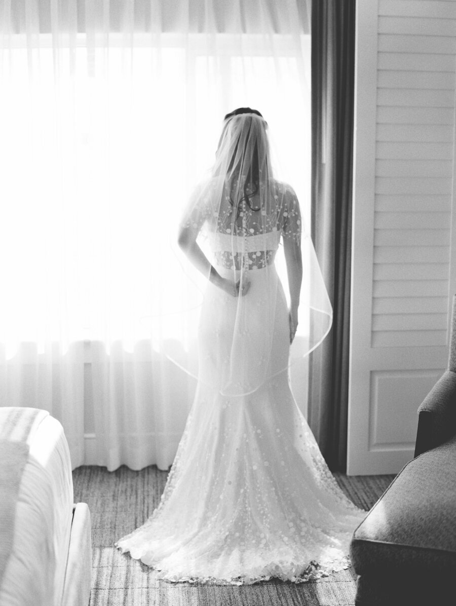 black and white photo of the back of a brides dress as she looks out the window