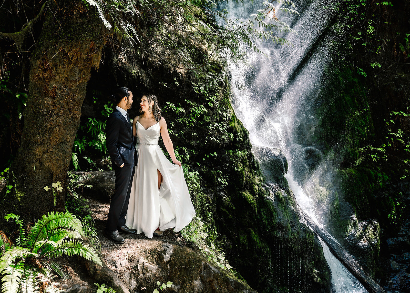 a bride and groom stand on the edge of a cliff. a waterfall streams behind them and light rays spill through the mist. they are surrounded by lush greenery as they smile sweetly at each other as their adventure elopement photographers document. 
