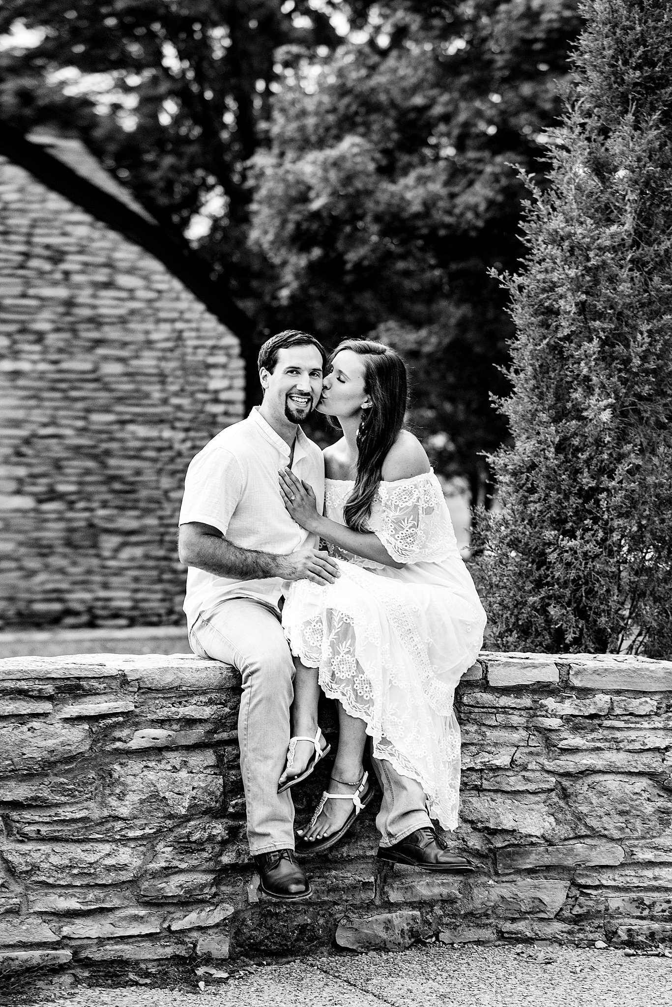 Black and White photo of couple sitting on rock wall outside of the Stone House at Arrington Vineyards, she is wearing a white lace dress and is leaning over to give her future husband a kiss on the cheek
