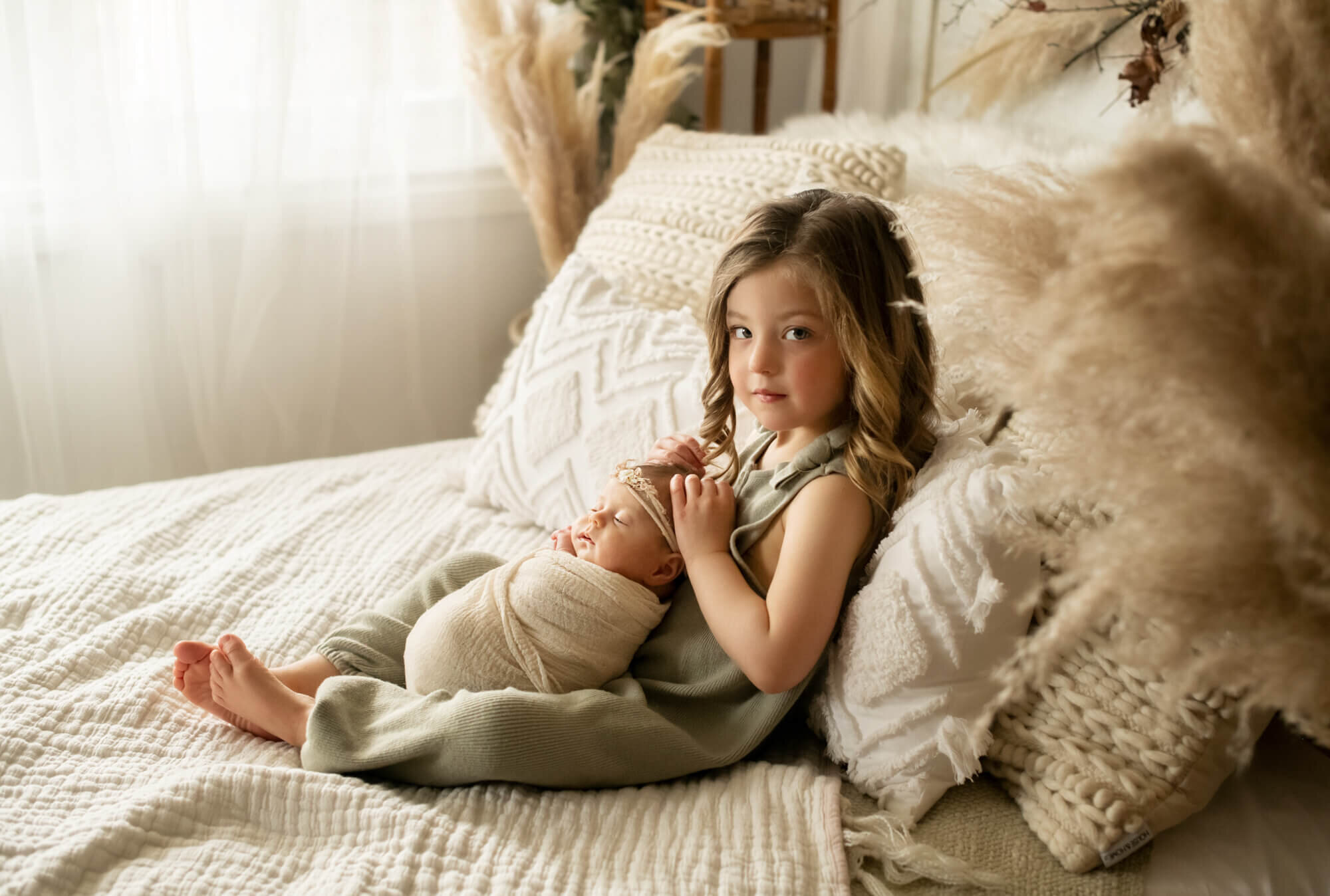 Older sister holding newborn baby sister in boho studio with pampas grass