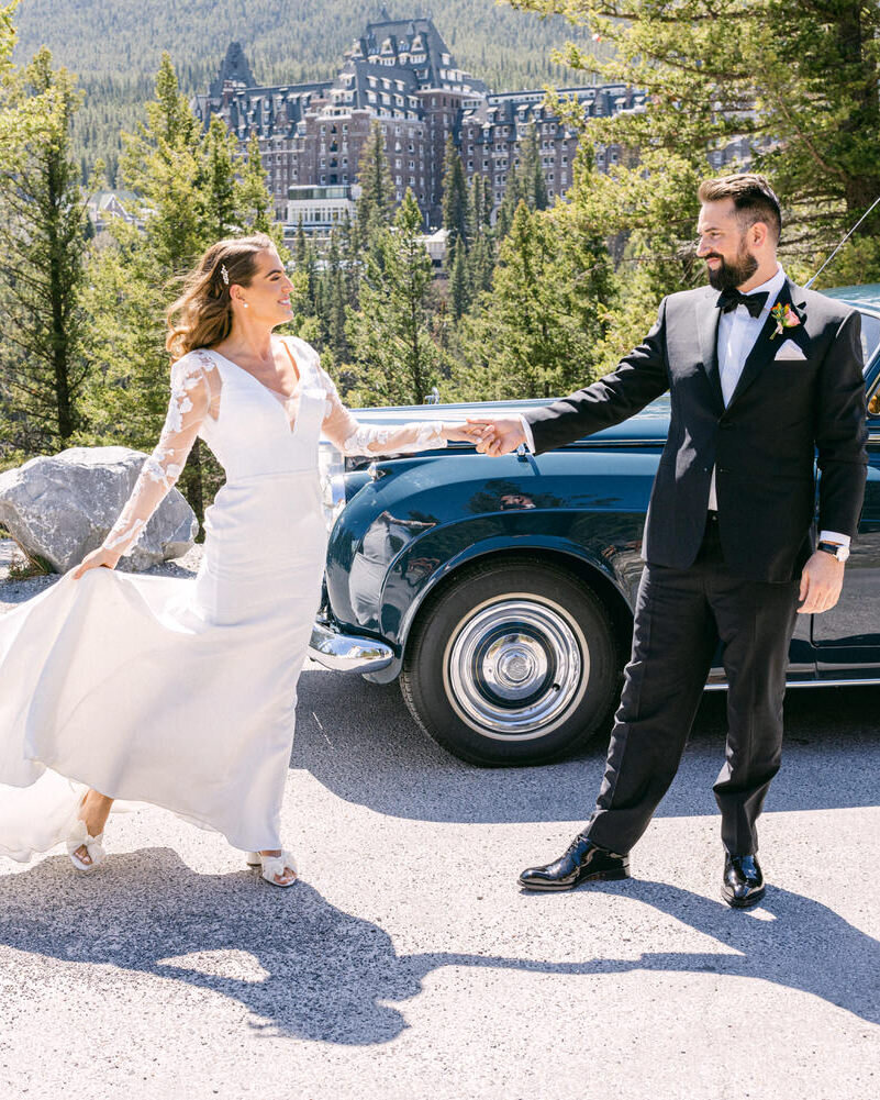This enchanting image captures the bride and groom dancing at Surprise Corner in Banff, with a luxury car parked behind them and the iconic Banff Springs Hotel in the background. The scene beautifully combines elegance and joy, set against the stunning backdrop of the hotel and natural landscape.