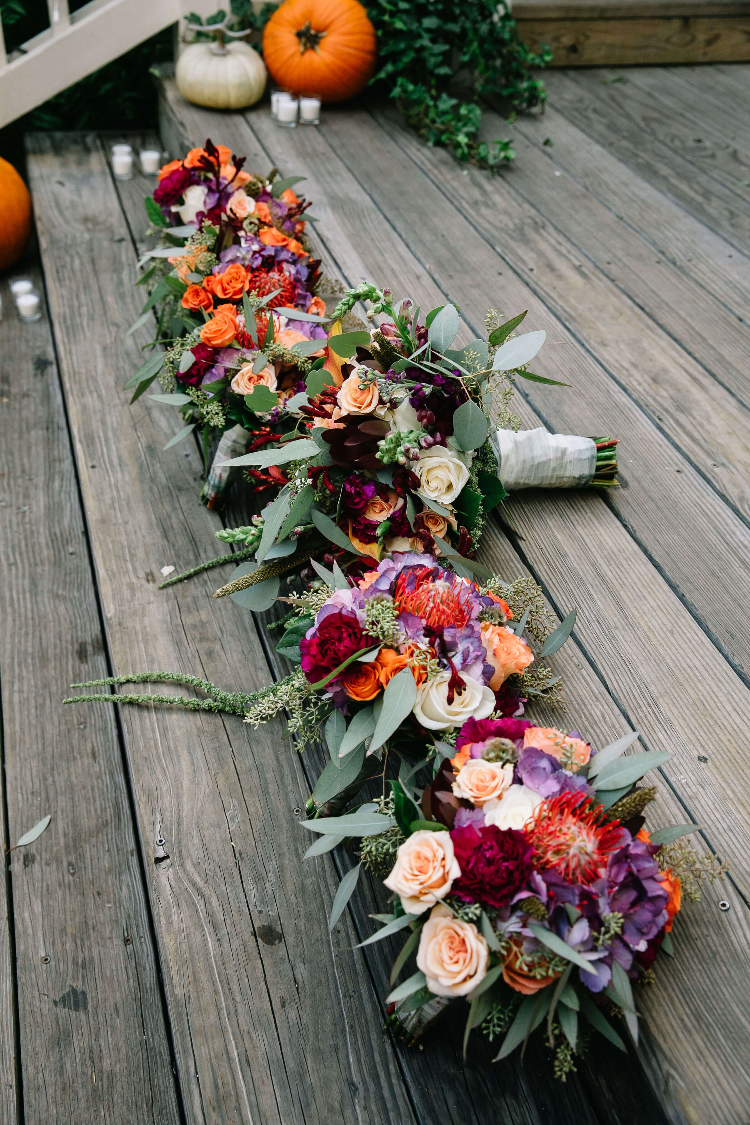 Several vibrant floral bouquets featuring roses, eucalyptus, and other colorful flowers are lined up on a wooden deck. Pumpkins and greenery are visible in the background, suggesting an autumn setting.