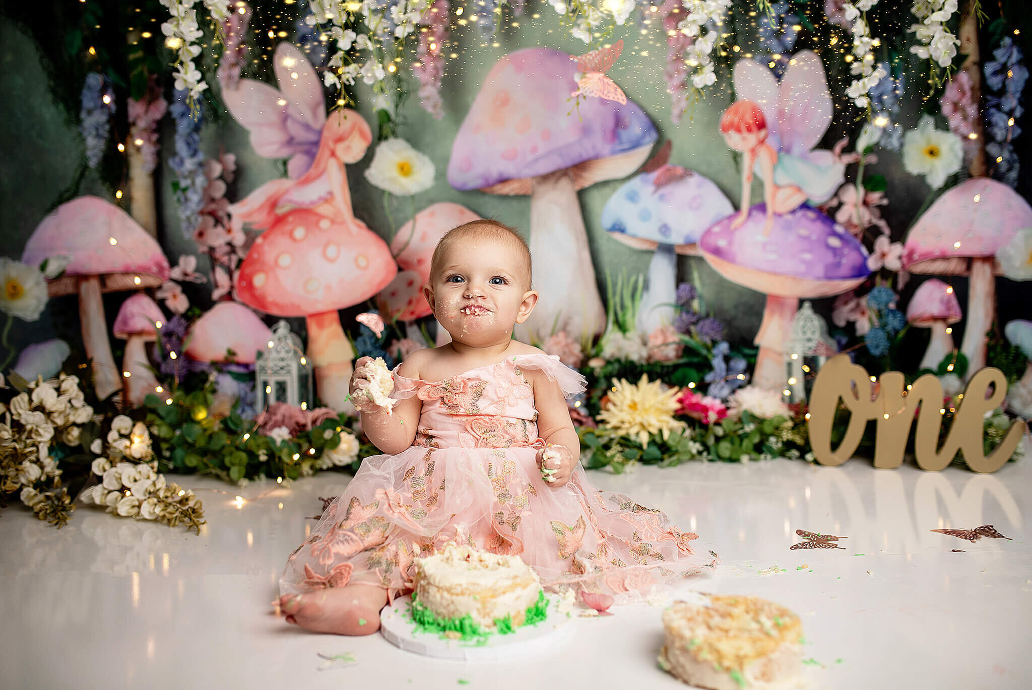 One year old cake smash with 1 year old girl sitting in front of a fairy tale background with mushrooms and fairy lights eating a cake