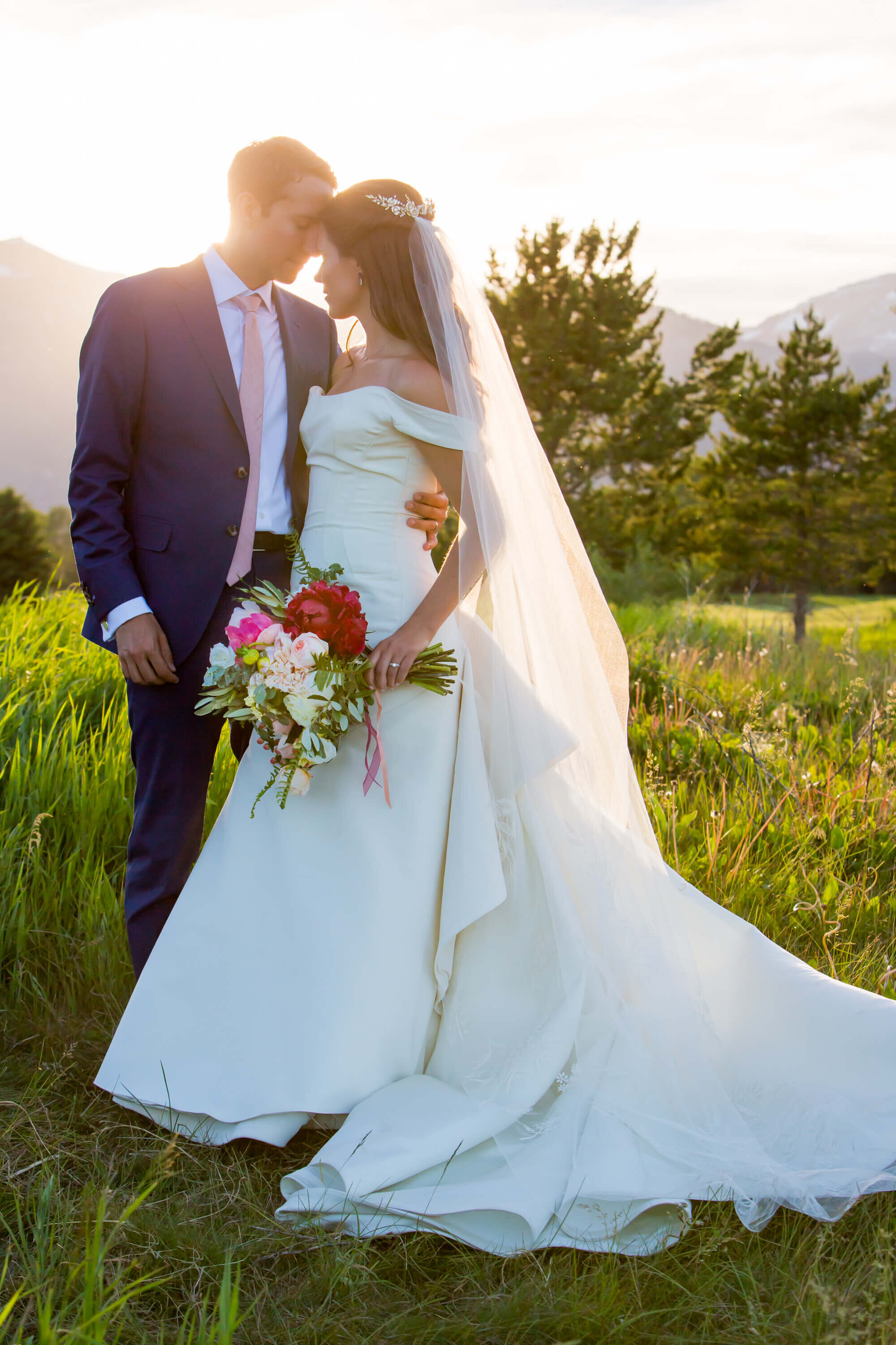 Bride in an off-the-shoulder gown and groom in a navy suit share a quiet moment at sunset at Jackson Hole Golf and Tennis Club, with soft golden light filtering through the Grand Teton landscape.