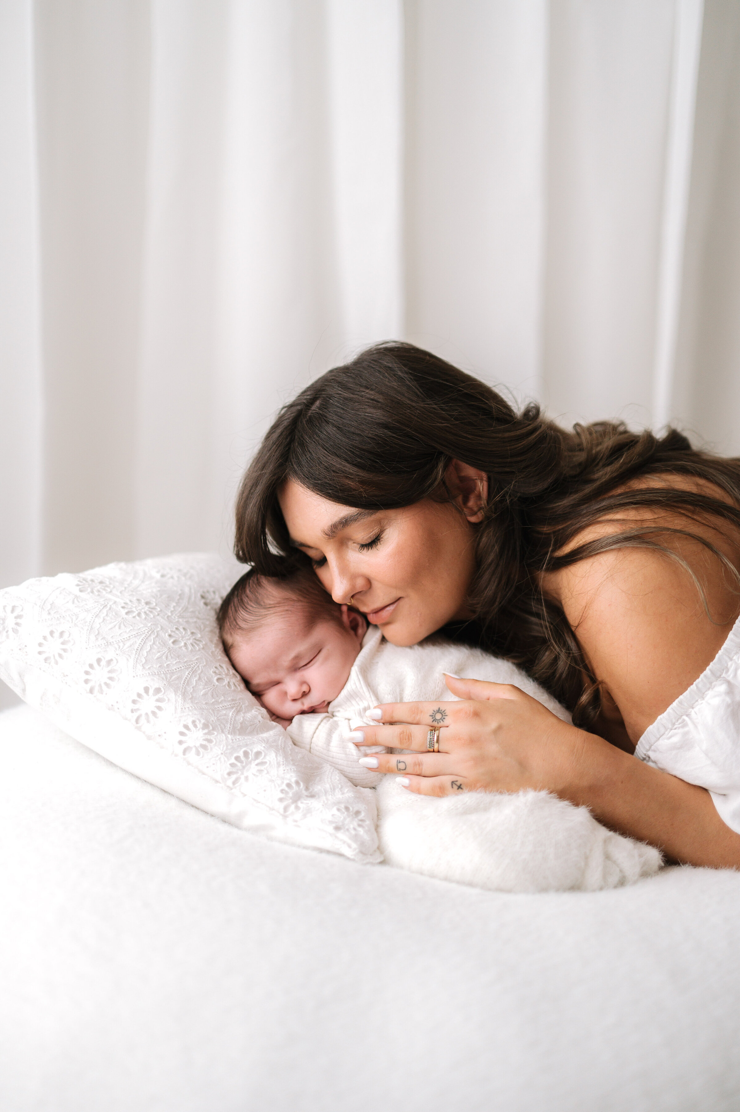 Baby girl sleeping on her side on a pillow and her mum leaning down and cuddling her at newborn photoshoot in Billingshurst