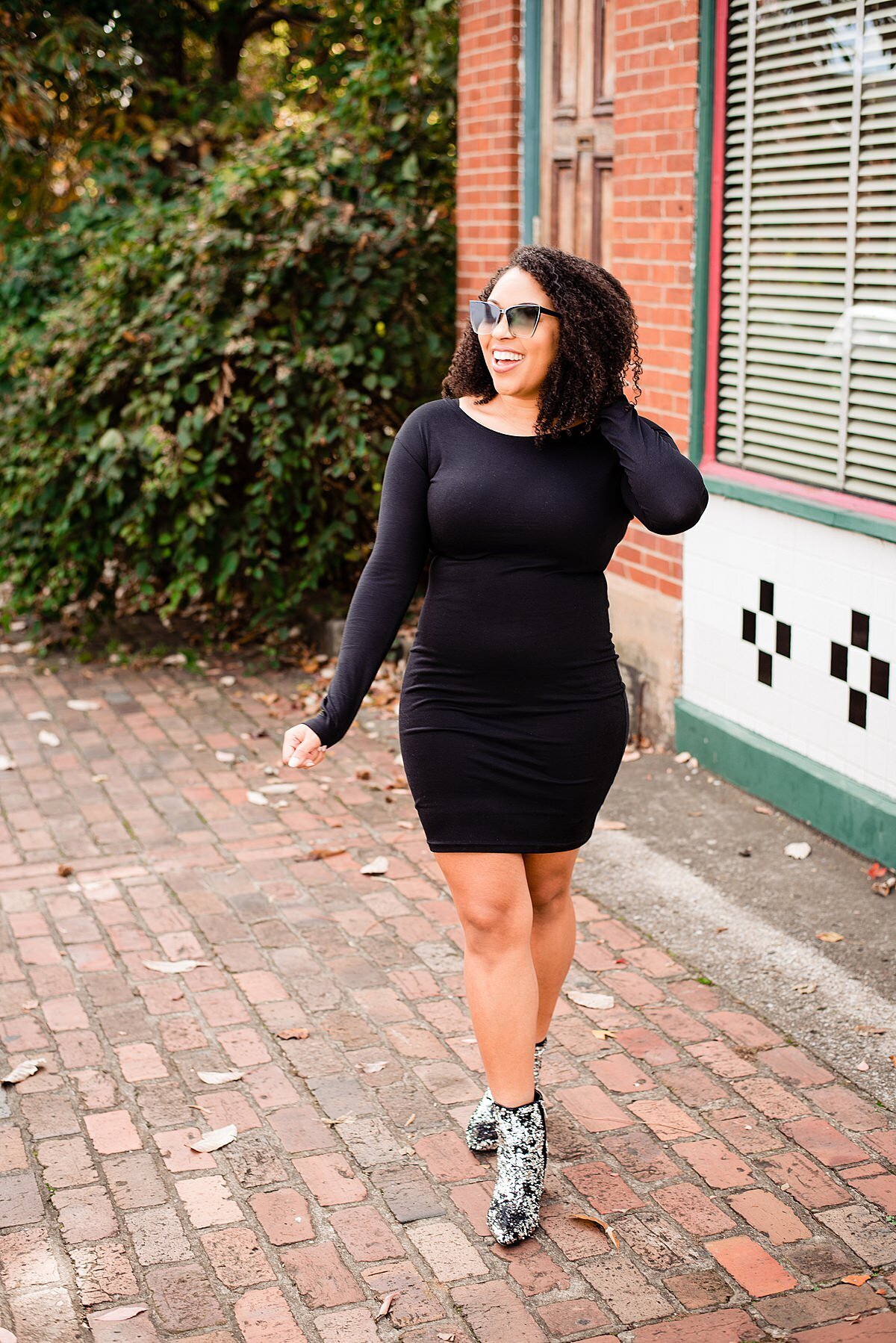 Jasmine Sweet strolling down sidewalk in Germantown wearing a fitted black dress, sequin boots and sunglasses