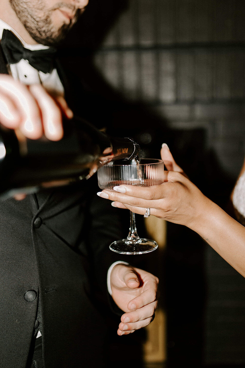 A close-up of a groom pouring champagne into a coupe glass held by the bride, highlighting her engagement ring. Captured at SoHo63, a Chandler wedding and event venue, this elegant moment reflects the luxury and celebration woven into every wedding hosted at the venue.