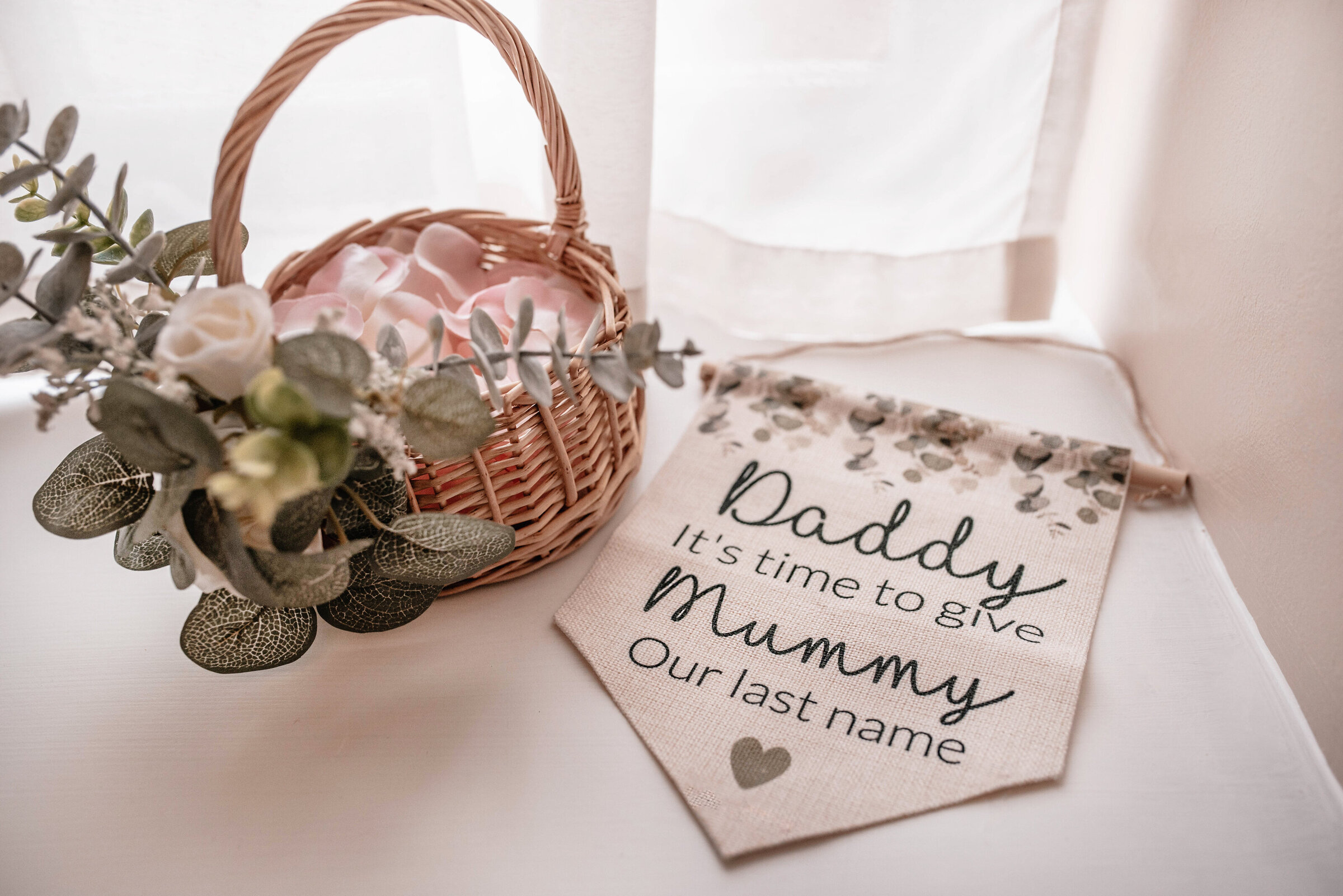 Wicker basket with floral arrangement containing petals seated on a windowsill next to woven page boy sign