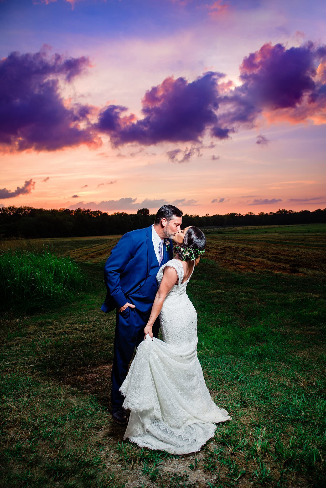 Bride holding her train and leaning into husband for a kiss during dramatic cloudy purple and pink sunset at Arrington Vineyards