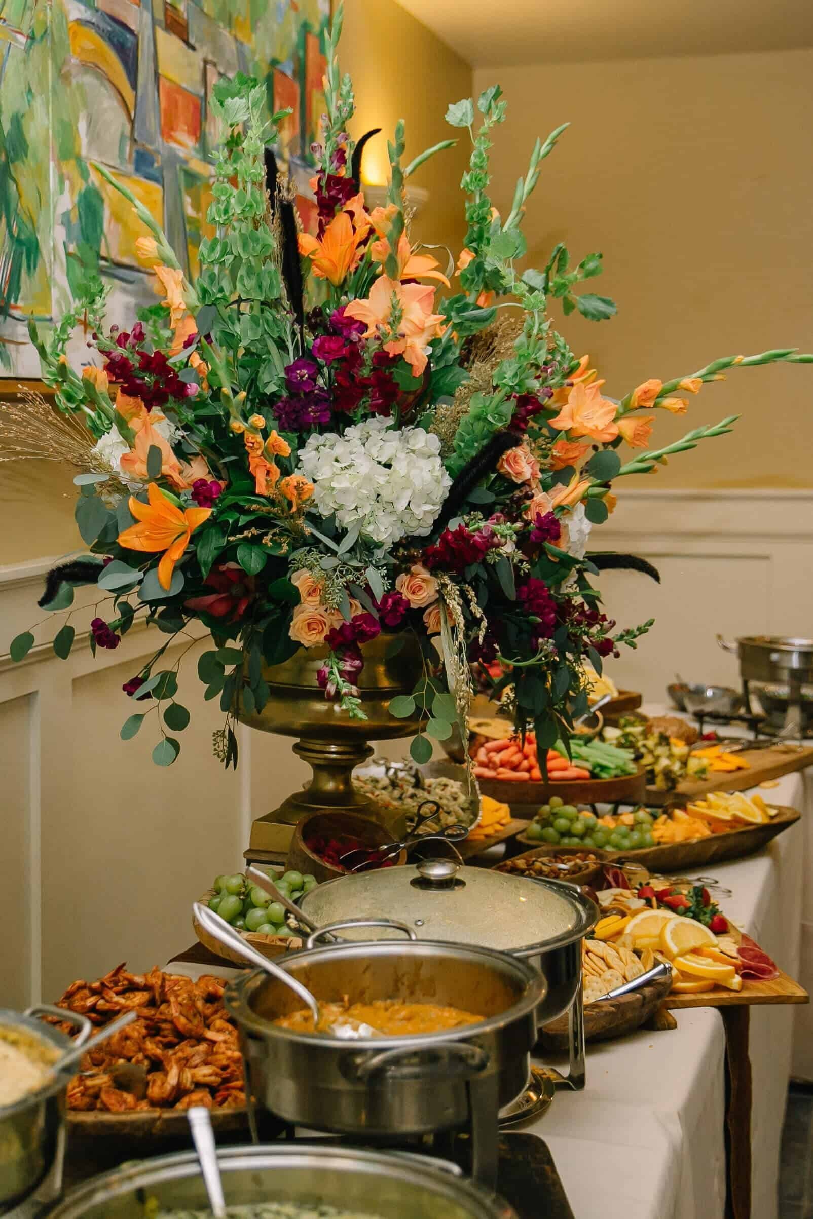 A buffet table with a wooden tiered stand holding assorted fresh fruits and leafy floral arrangements, surrounded by cheese, crackers, vegetables, and a tray of sandwiches in a bright, elegant room.